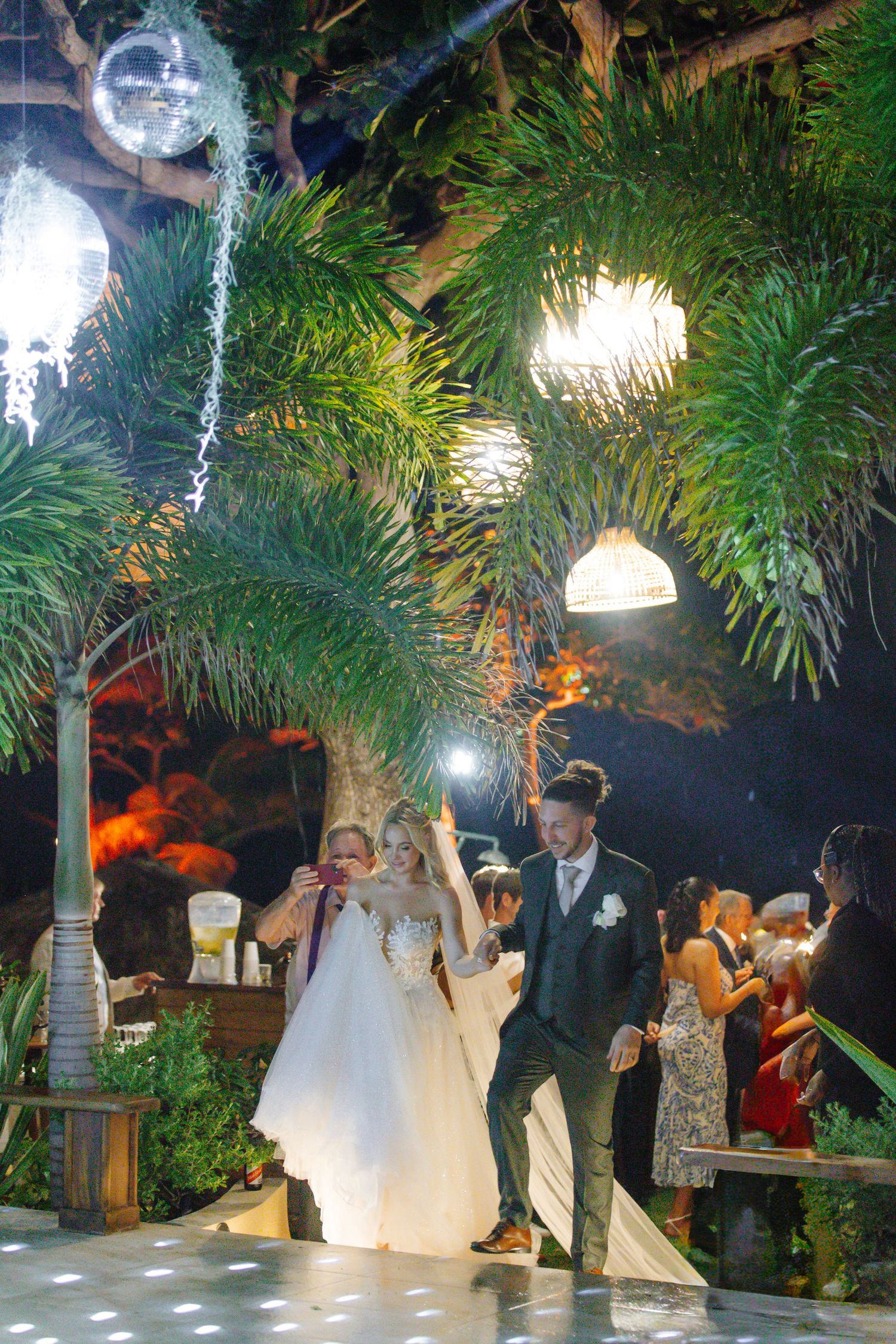 Bride and groom dancing joyfully at tropical wedding reception with palm trees, disco ball, and sparkling lights.