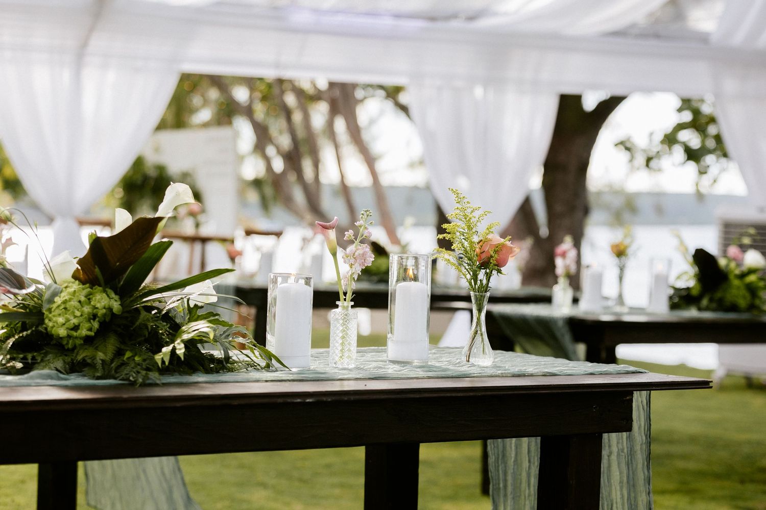 Elegant outdoor wedding reception table with tropical floral arrangements, white candles, and draped tent backdrop.