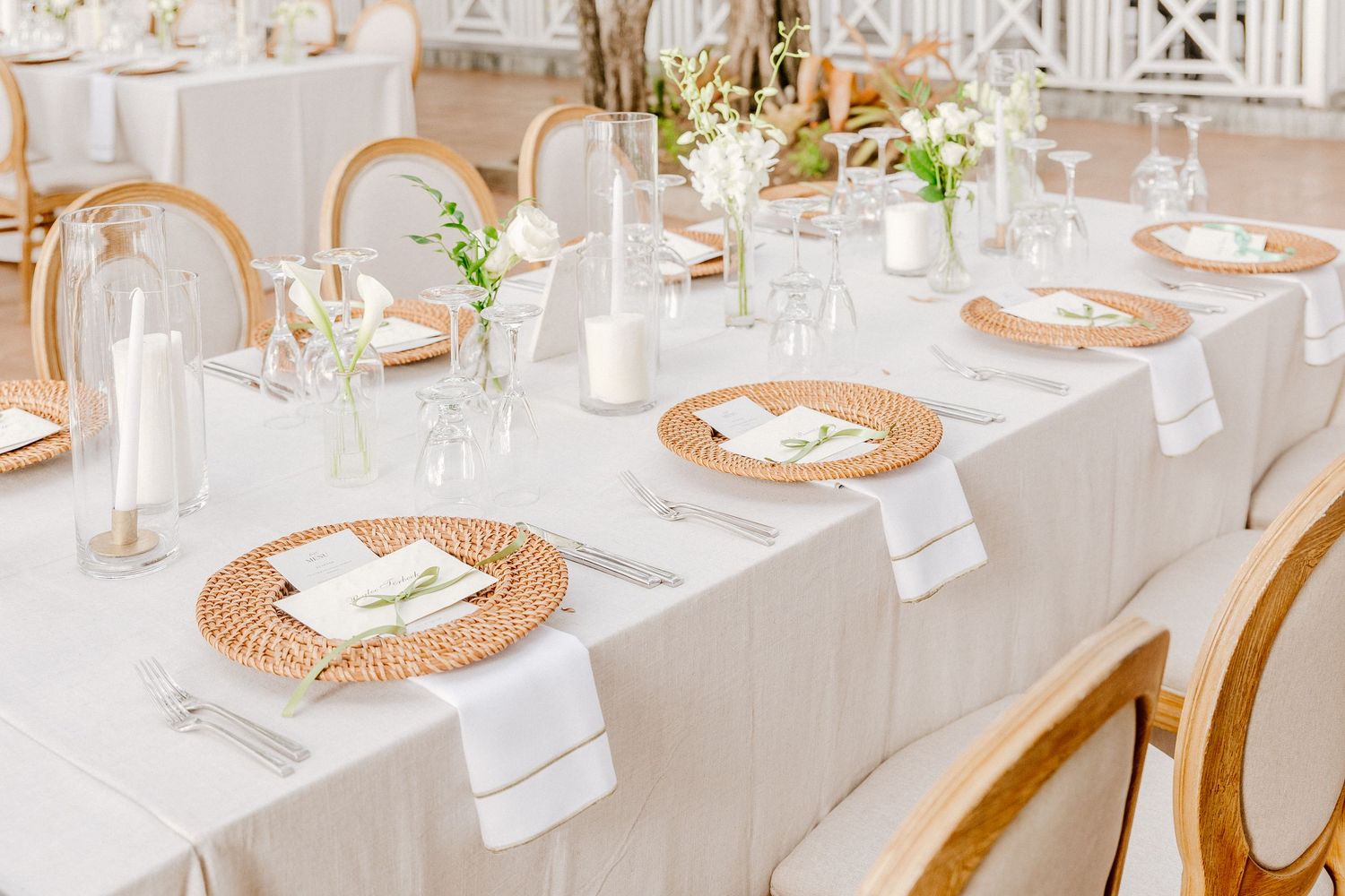 Elegant wedding reception table with woven chargers, white linens, candles, and floral centerpieces in a bright venue.