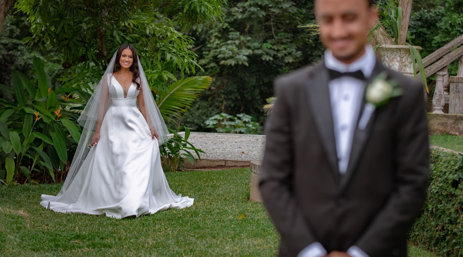 Bride in white gown and veil walking toward groom in black tuxedo in lush garden setting.