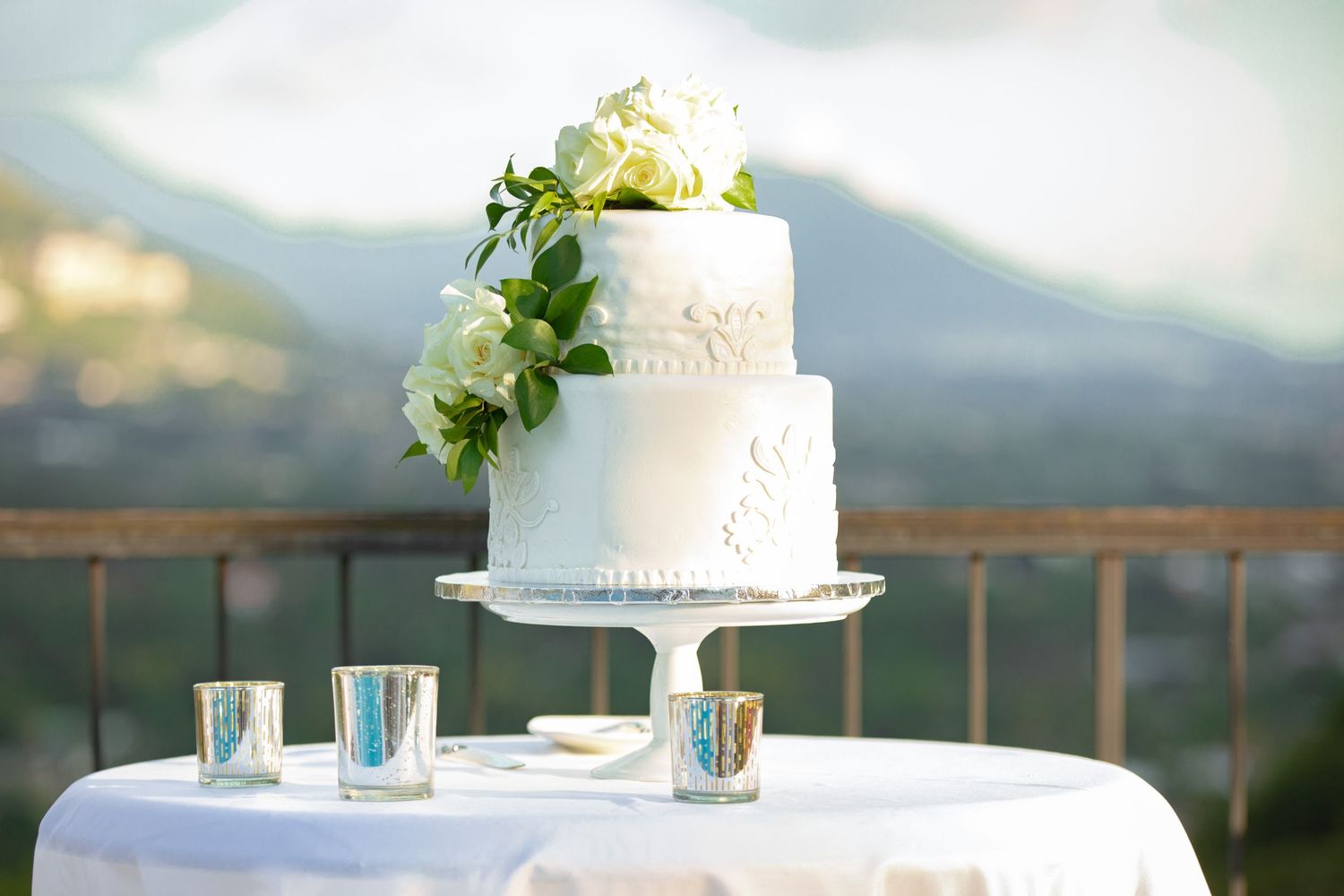 Two-tier white wedding cake decorated with white roses and greenery on a outdoor table with mountain backdrop.