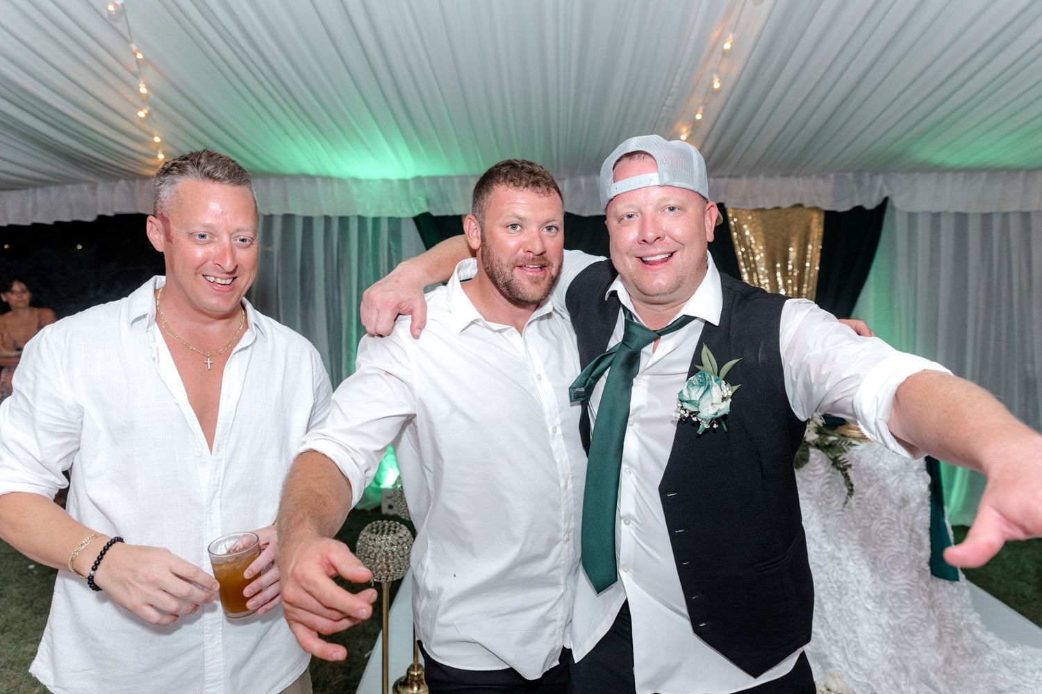 Three smiling men posing together at a wedding reception under a white tent.