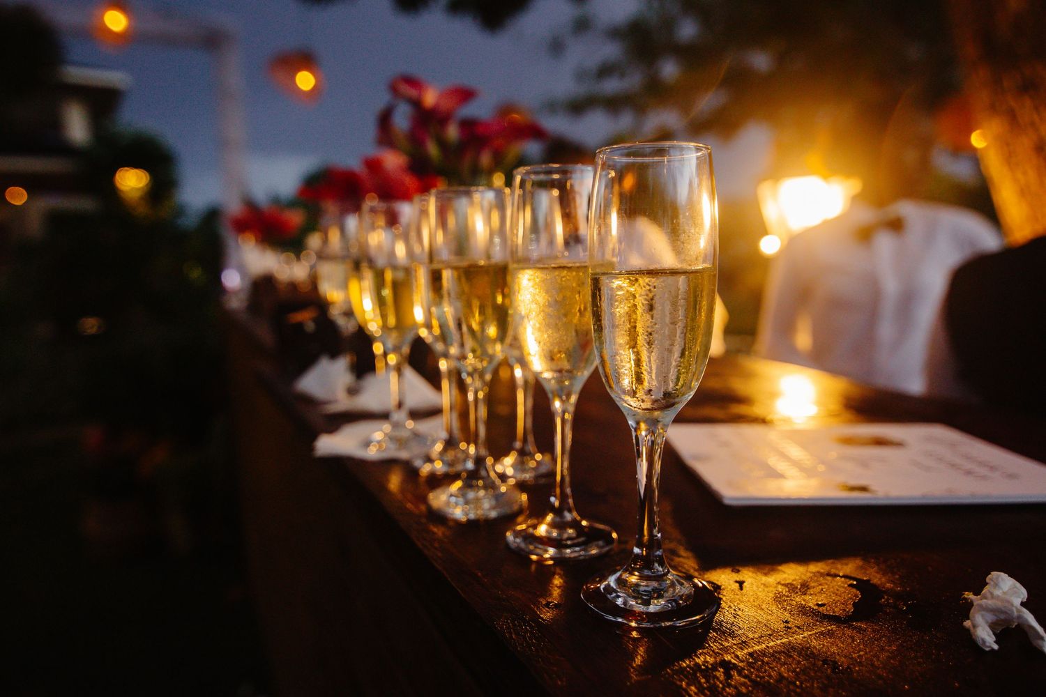 Row of elegant champagne flutes filled with sparkling wine at an outdoor evening reception with warm glowing lights.