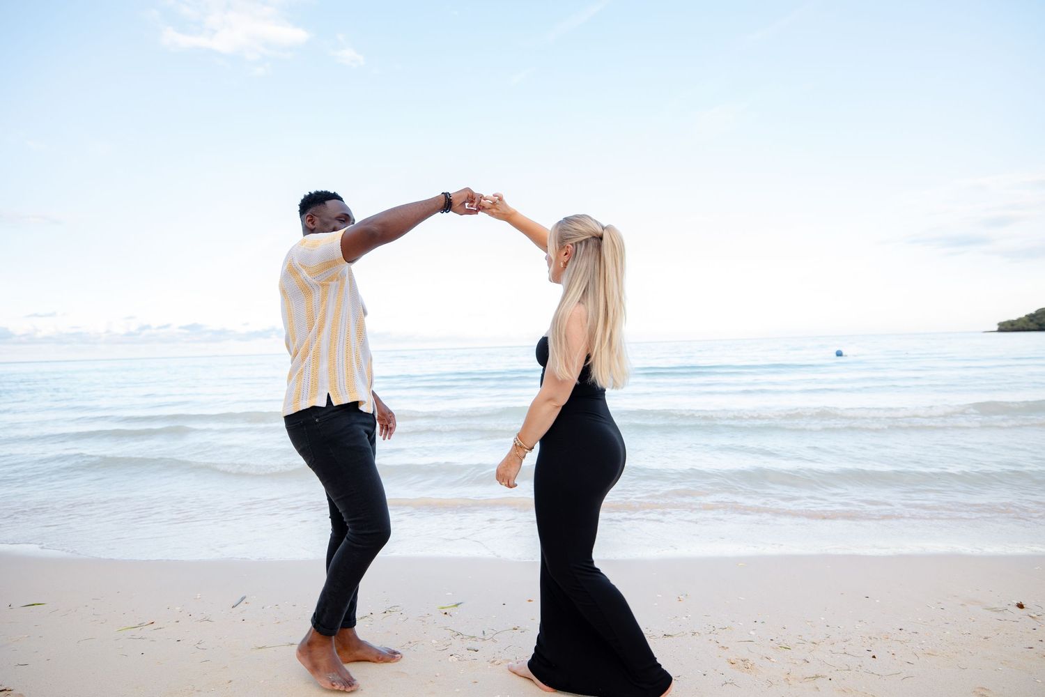 Couple dancing on a tropical beach, man spinning woman in black maxi dress near ocean waves.