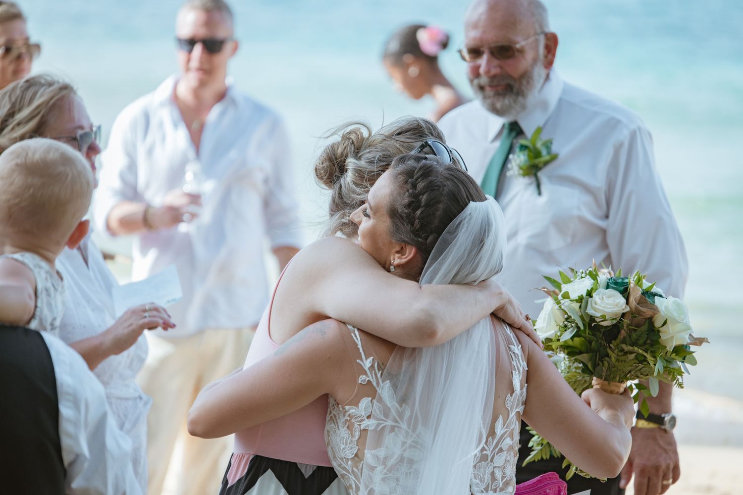Bride hugging a guest at a beach wedding ceremony while holding a white bouquet.