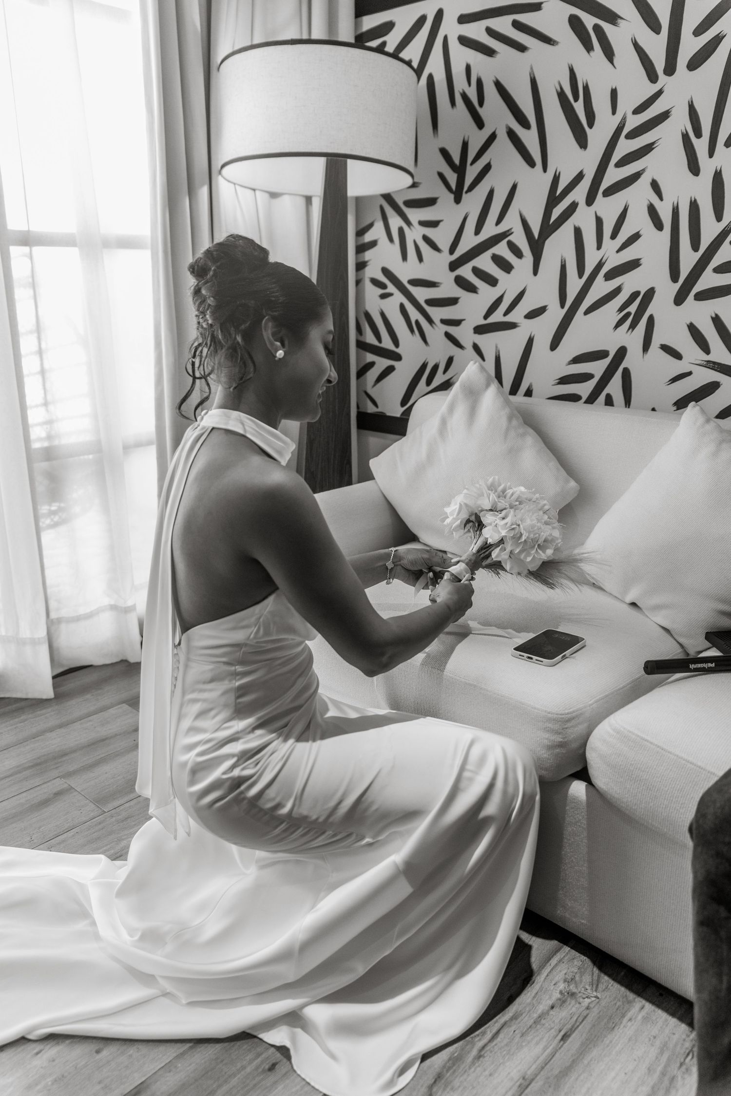 Black and white photo of bride in halter wedding gown sitting on sofa holding bouquet in hotel room.