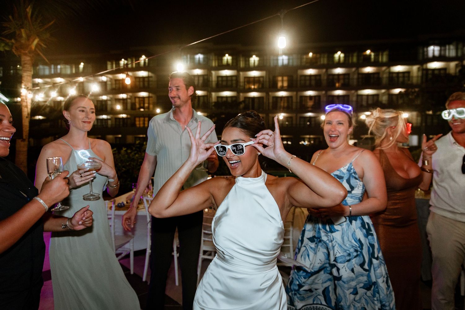 Bride dancing with sunglasses and guests at outdoor nighttime beach wedding reception.