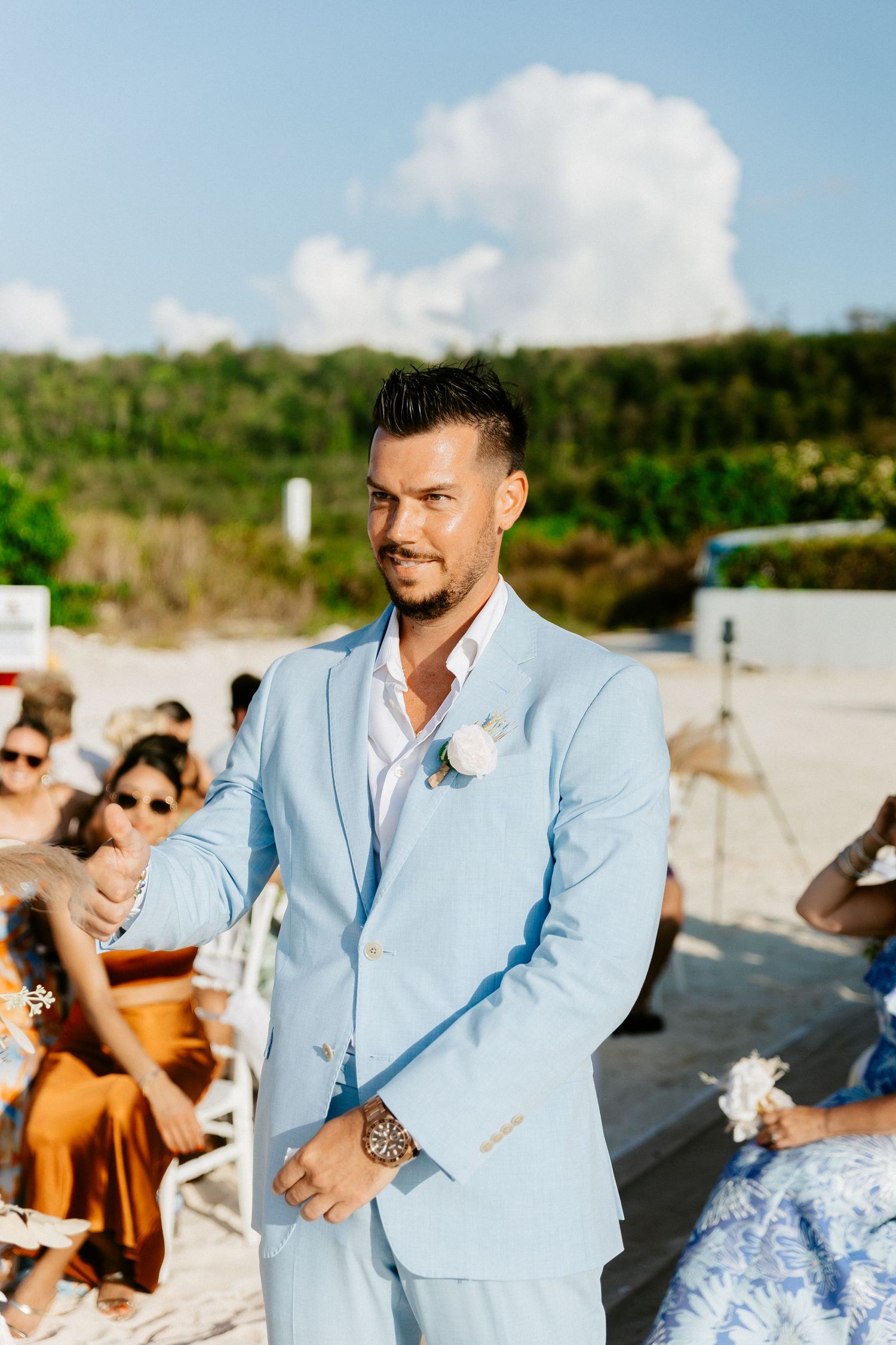 Groom in a light blue suit with boutonniere stands at outdoor beach wedding ceremony aisle.