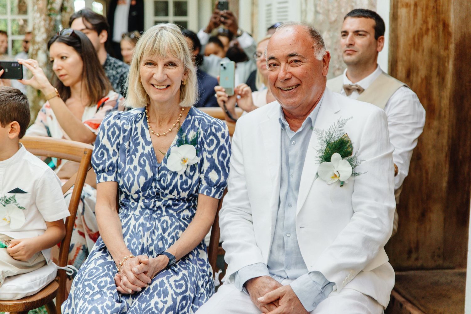 Smiling couple in elegant wedding attire seated at an outdoor ceremony, man in white suit, woman in blue floral dress.