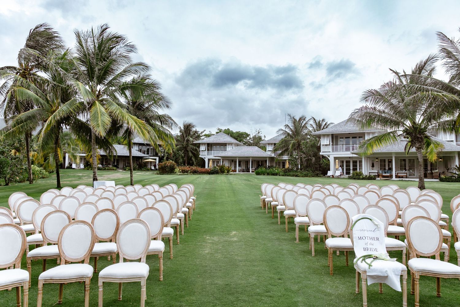 Elegant outdoor wedding ceremony setup with white chairs on green lawn, palm trees, and white colonial buildings.