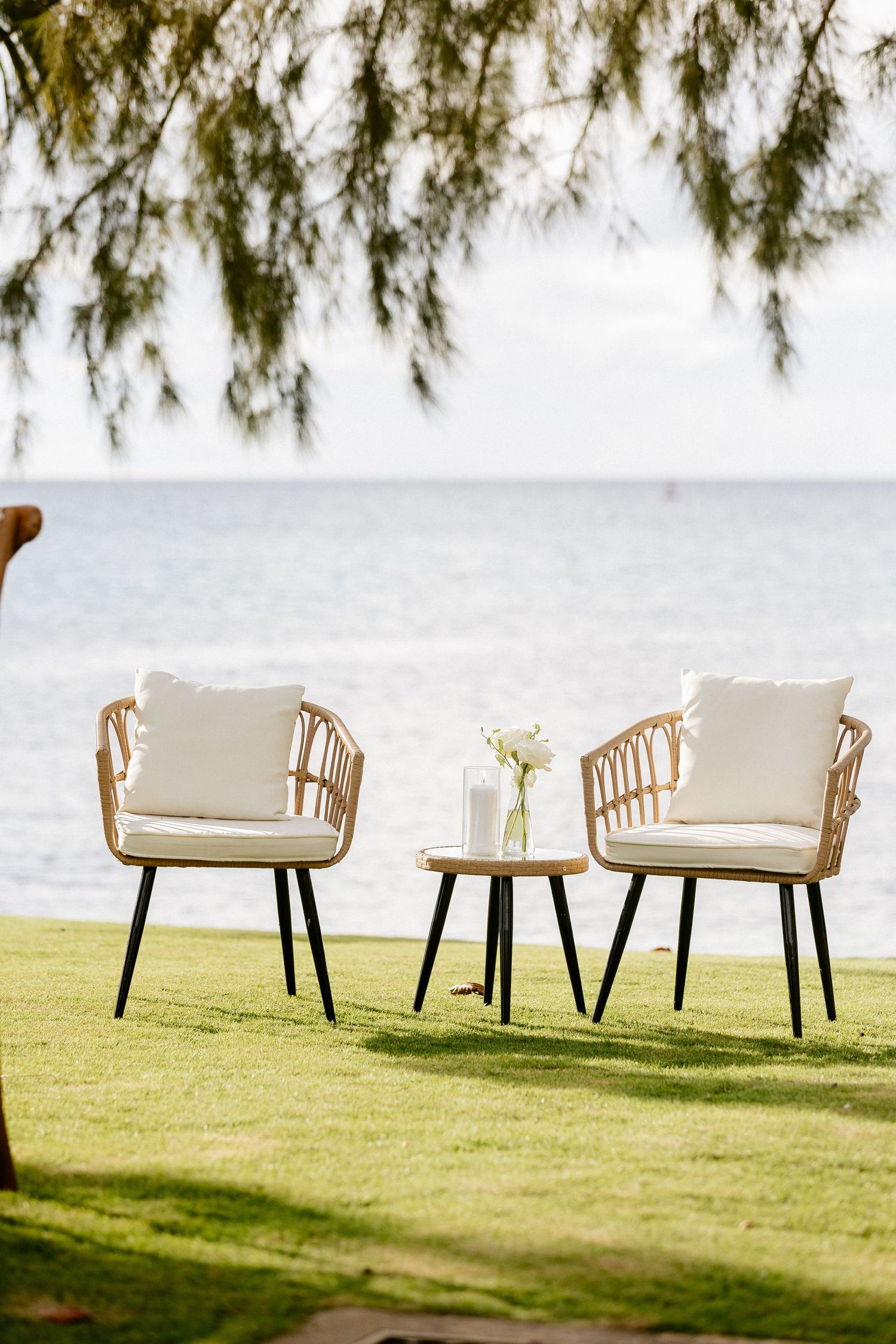 Two rattan chairs with white cushions and a small table set on green lawn overlooking a calm ocean view.