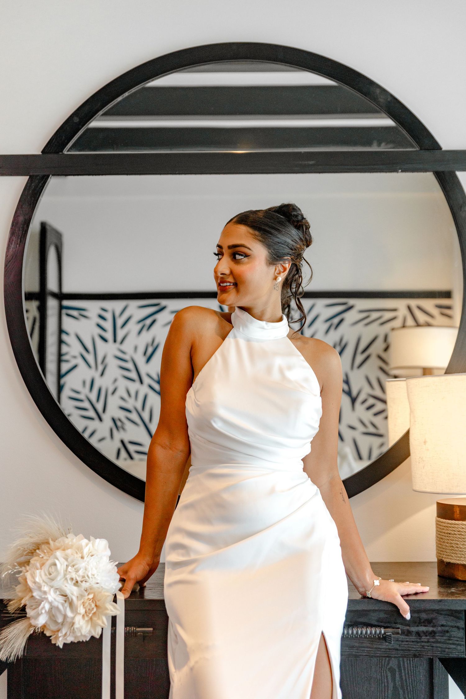 Elegant bride in a white halter gown posing in front of a large round mirror holding a bouquet.