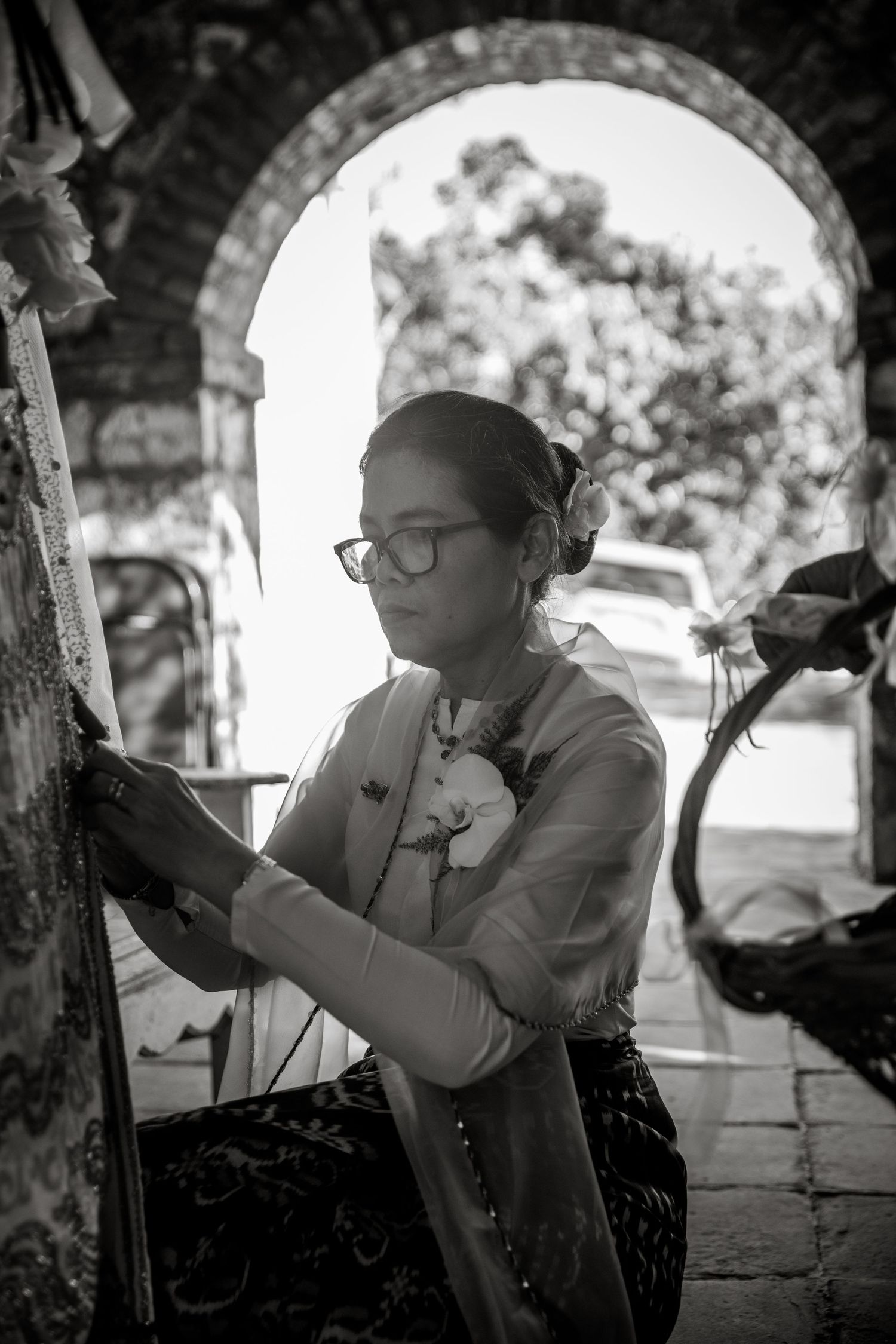 Black and white photo of young woman with glasses adjusting wedding decorations under stone arch.