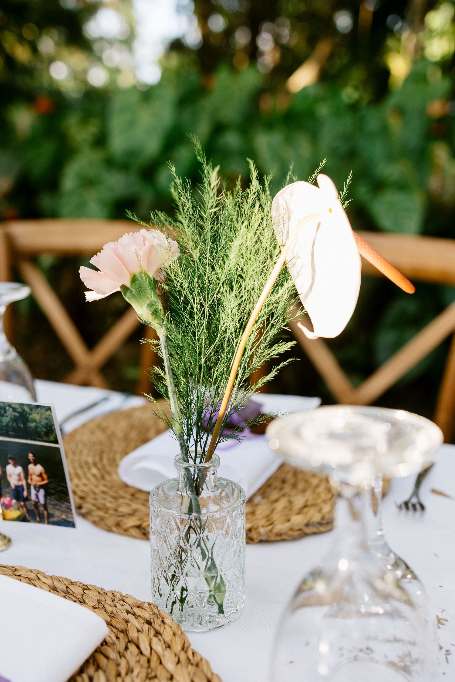 Glass vase with pink carnation and greenery centerpiece on a white outdoor wedding reception table.