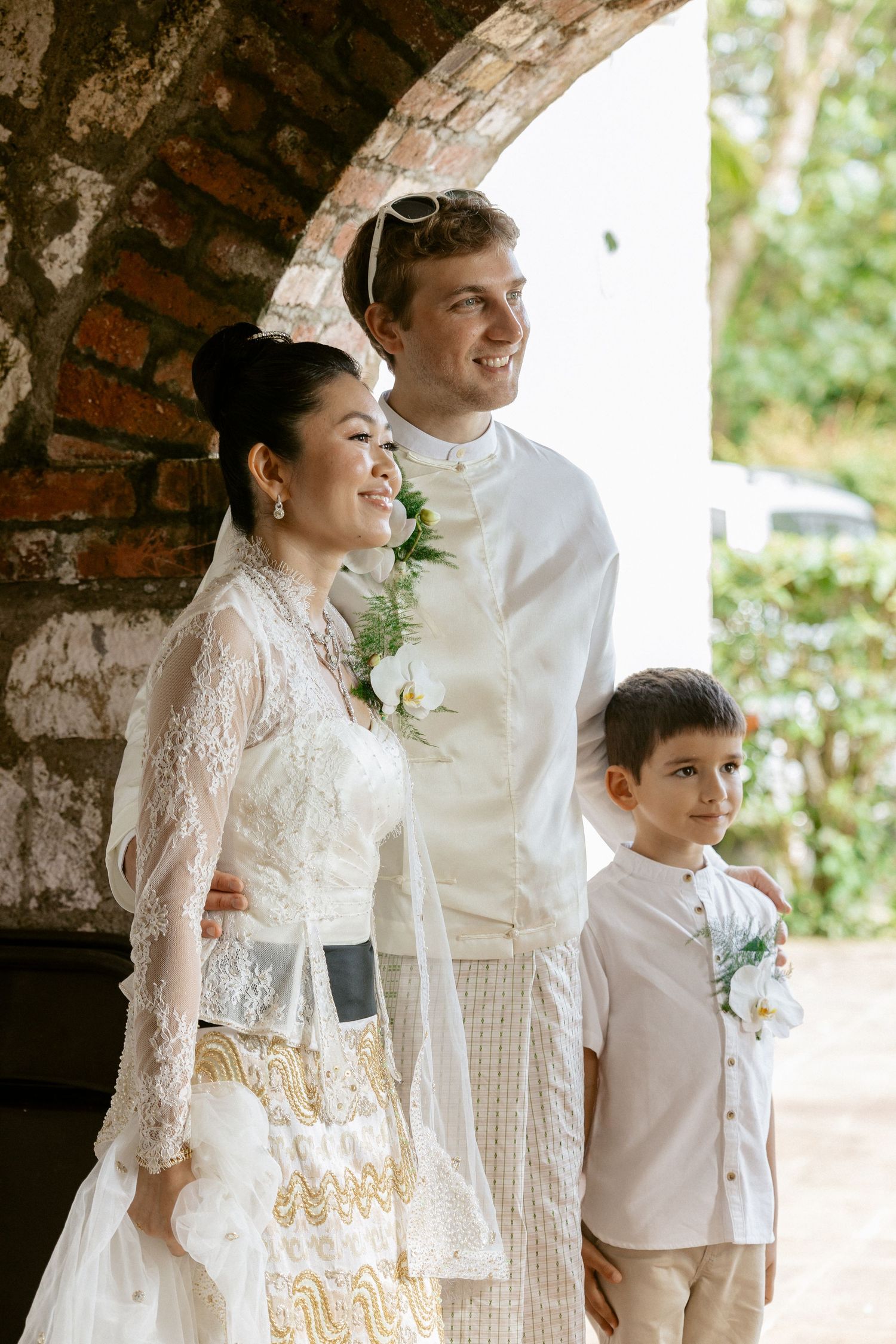 Bride and groom in traditional white wedding attire stand with young boy under rustic brick arch.