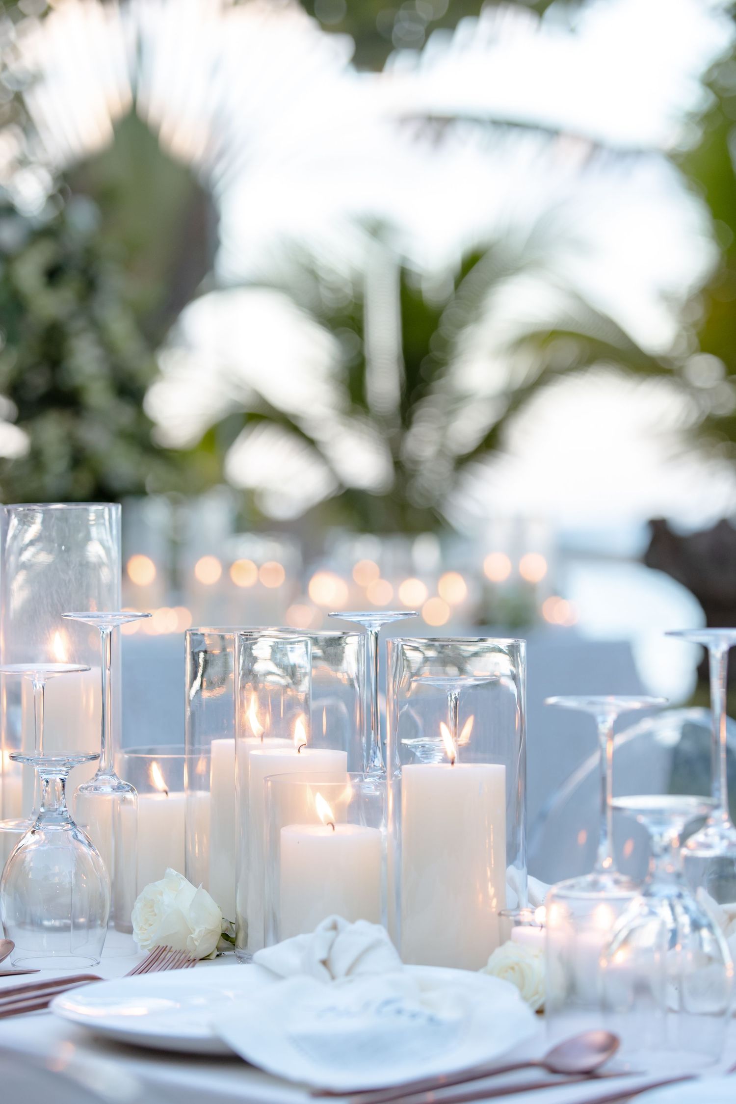 Elegant outdoor wedding table with white pillar candles in glass holders, wine glasses, and tropical palm trees in background.