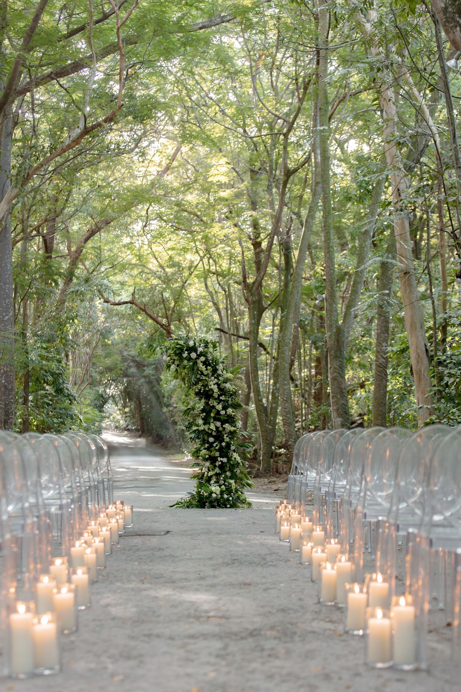 Outdoor wedding ceremony aisle lined with white pillar candles and ghost chairs under a lush green tree canopy.