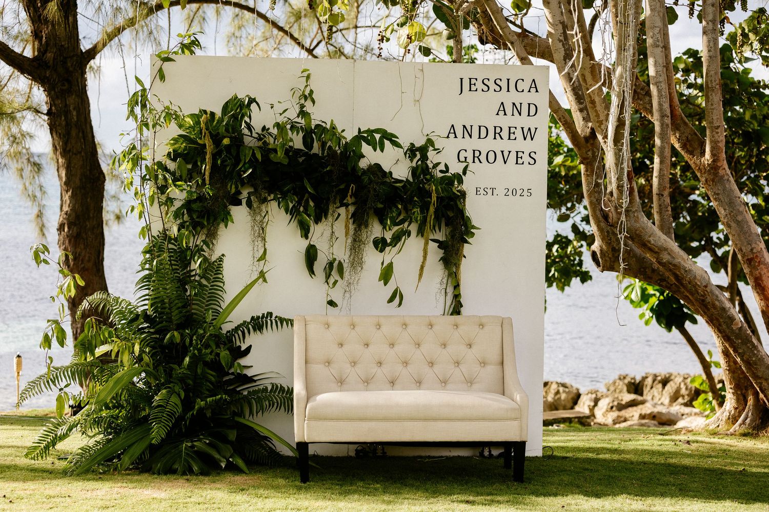 Elegant outdoor wedding backdrop with white tufted loveseat, lush greenery, and Jessica and Andrew Groves Est. 2025 sign.