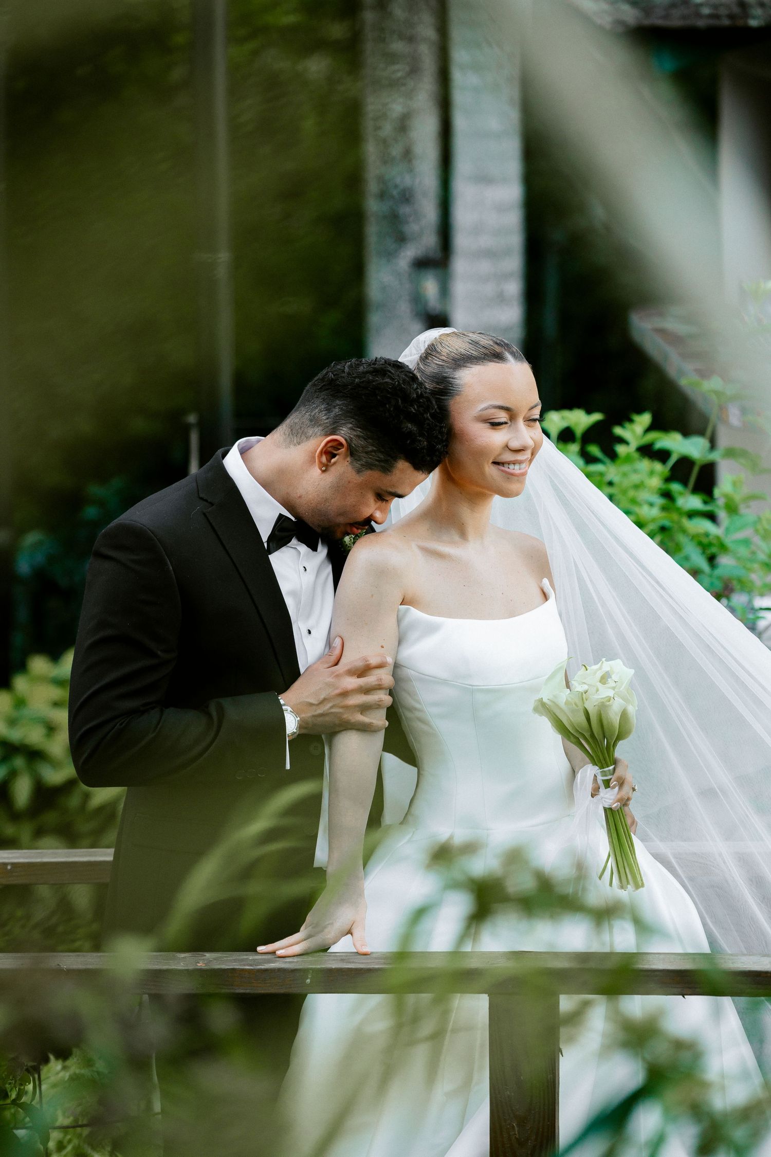 Groom in black tuxedo tenderly kisses brides shoulder as she holds white bouquet outdoors.