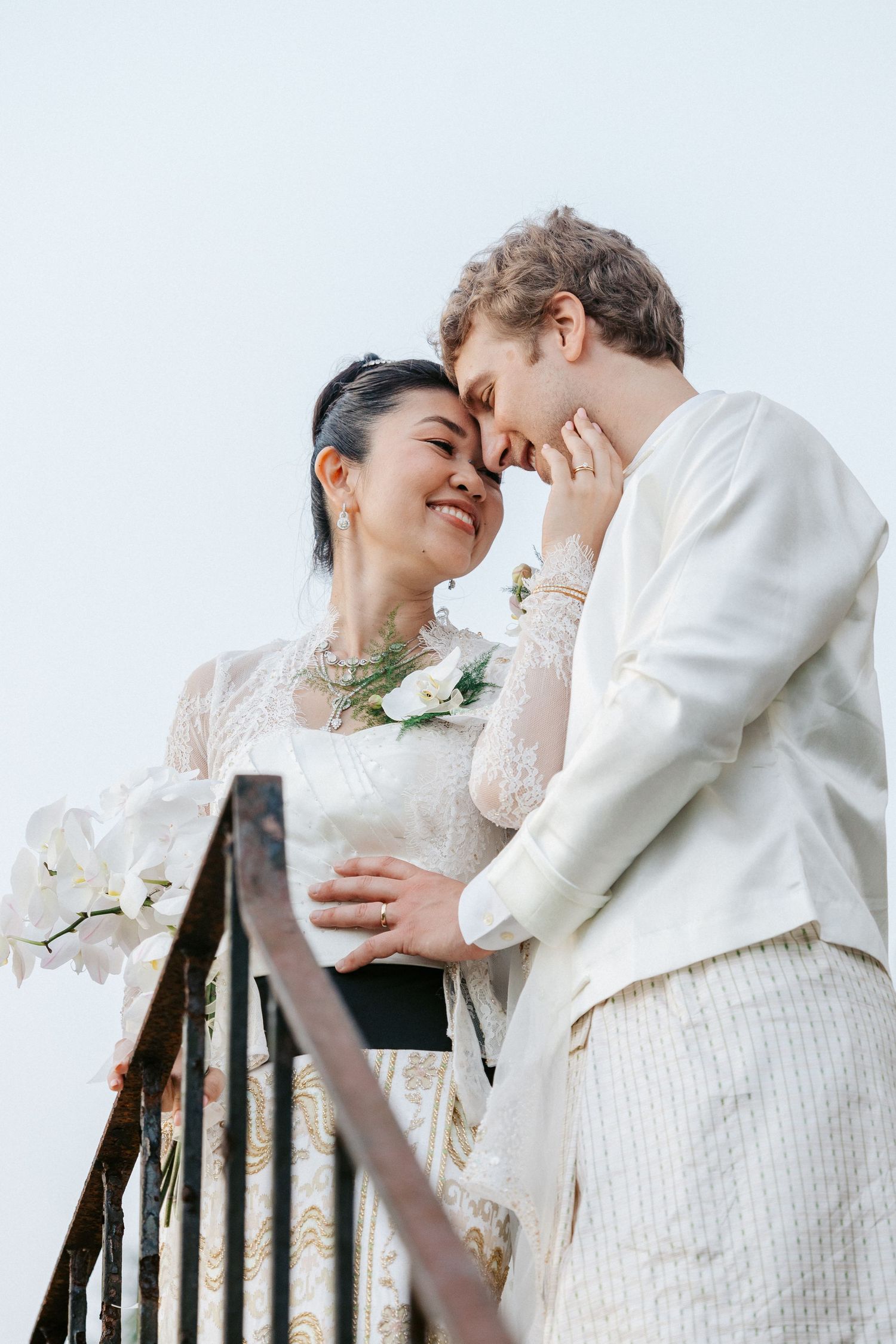 A joyful interracial couple in white wedding attire share a tender moment on a balcony adorned with orchids.