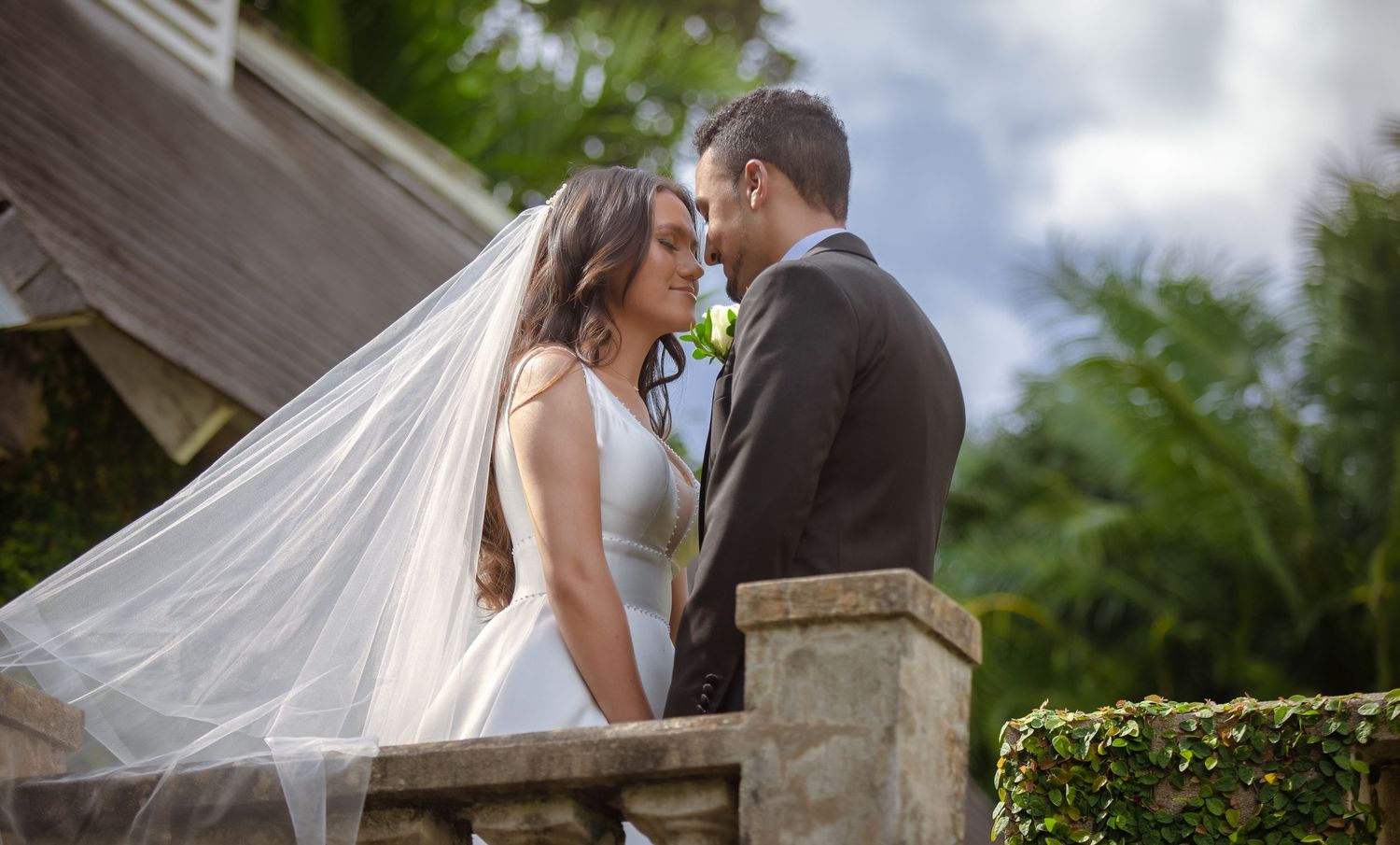 Bride and groom share romantic moment on stone balcony with veil flowing in the breeze.