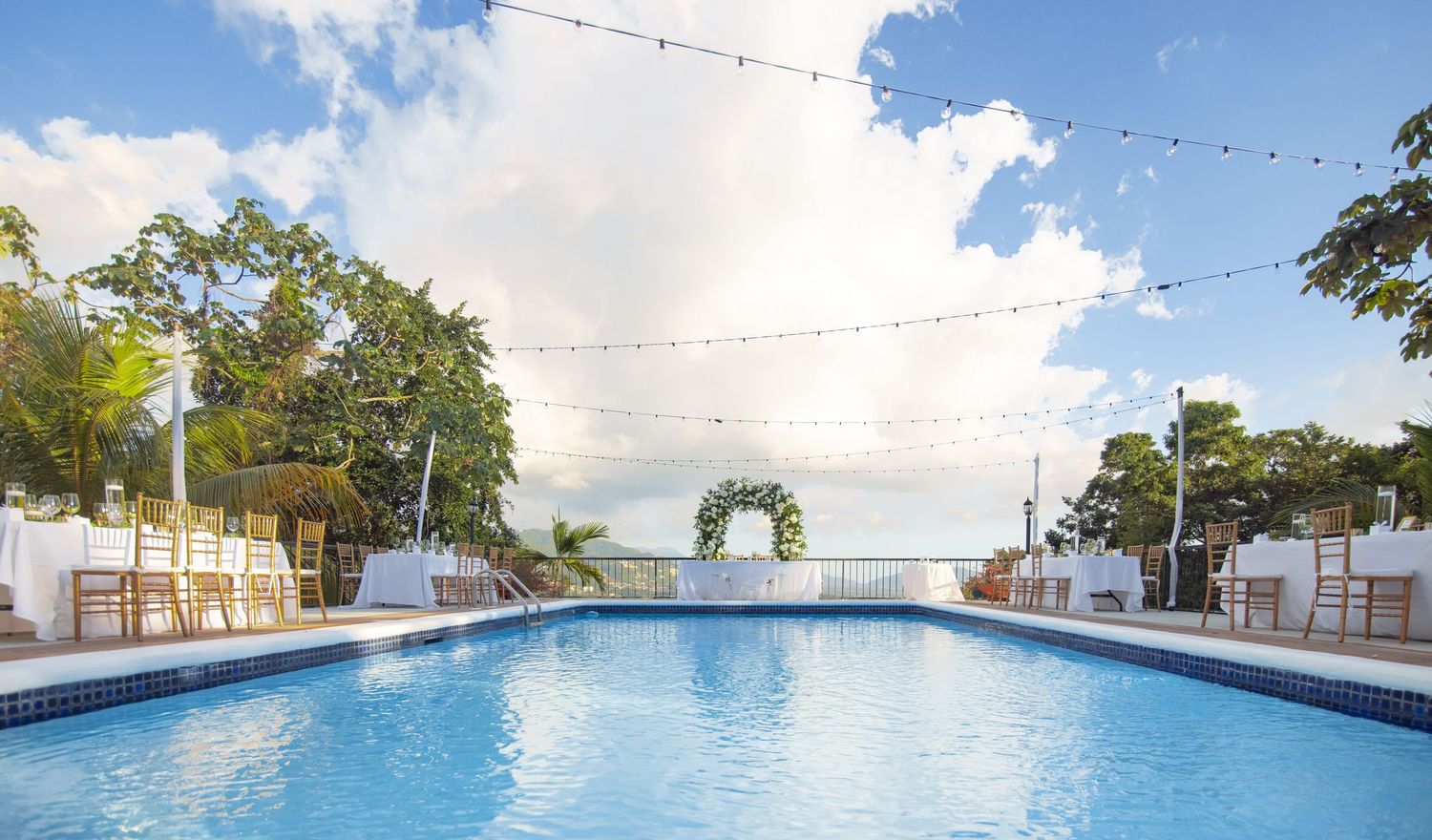 Stunning outdoor wedding venue featuring a blue pool, string lights, and floral arch with ocean views.