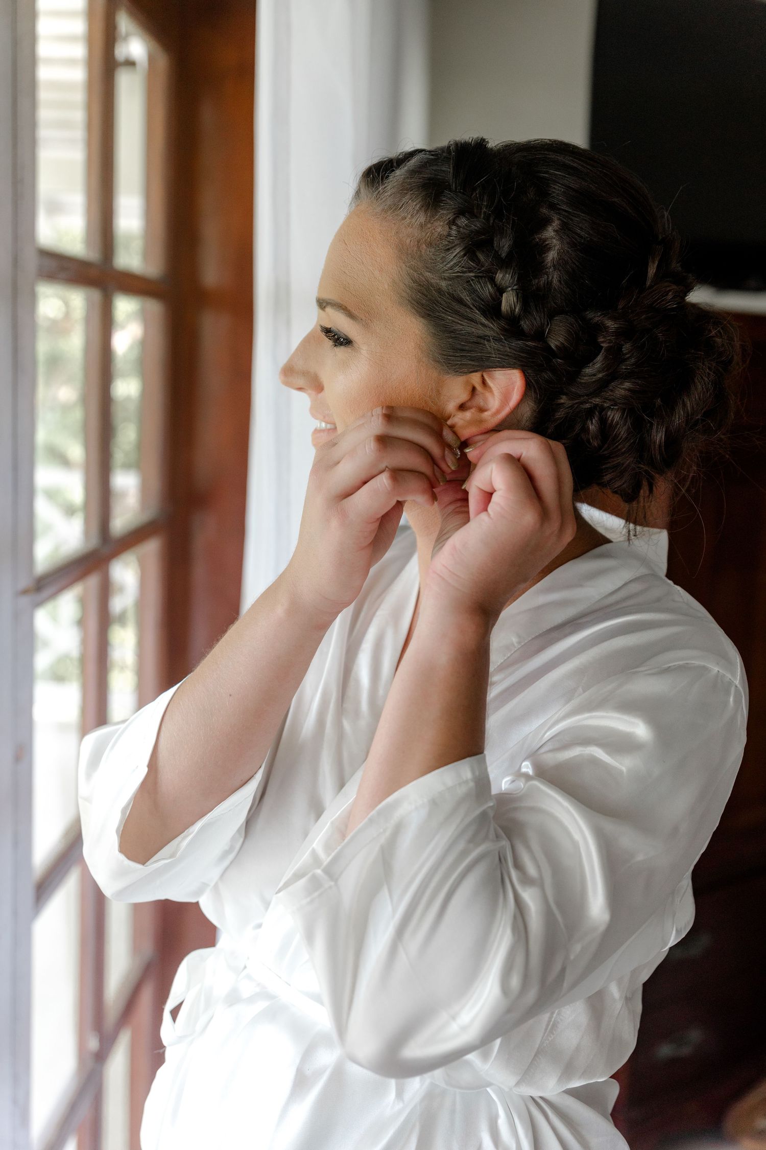Bride in white satin robe with braided updo putting on earrings by a sunlit window.