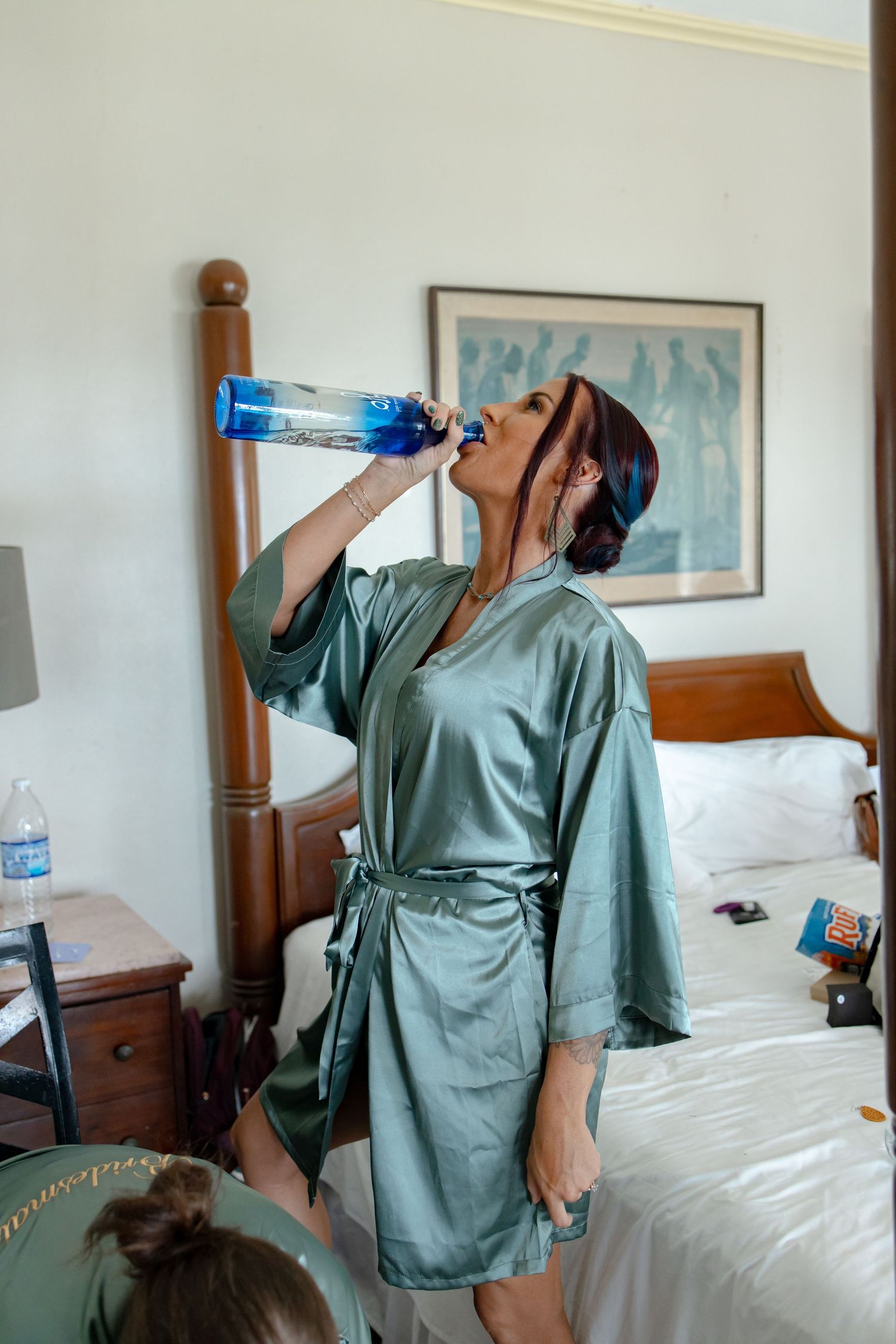 Bridesmaid in sage green robe drinking water bottle while getting ready.