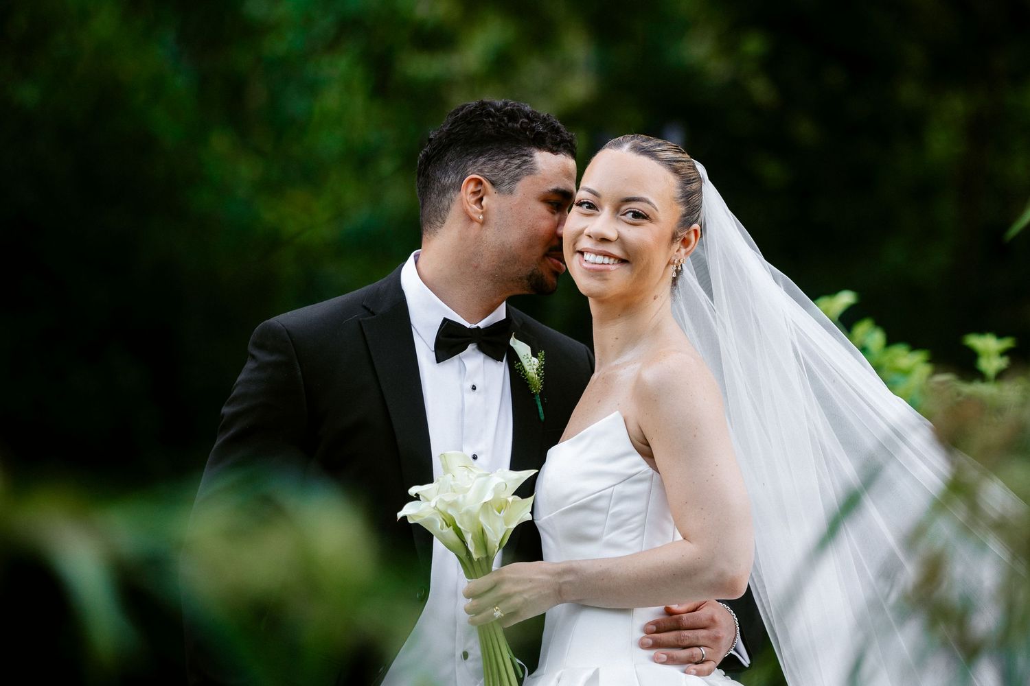 Bride smiles joyfully as groom kisses her cheek during outdoor wedding portrait surrounded by lush greenery.