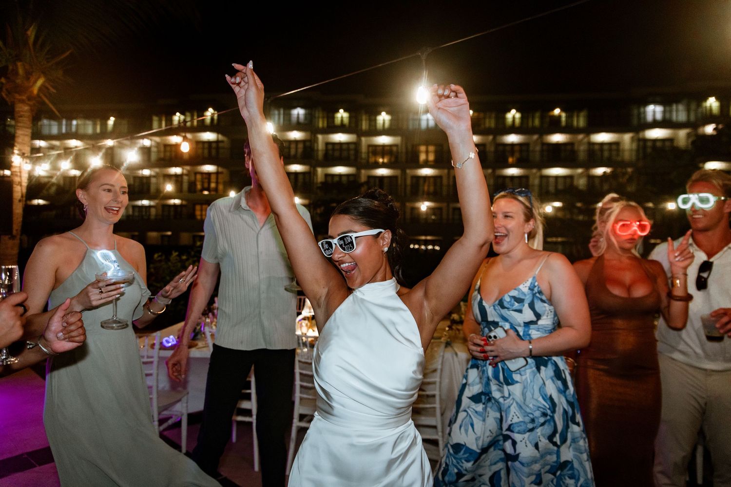 Guests dancing and celebrating at an outdoor wedding reception at night with colorful lights.