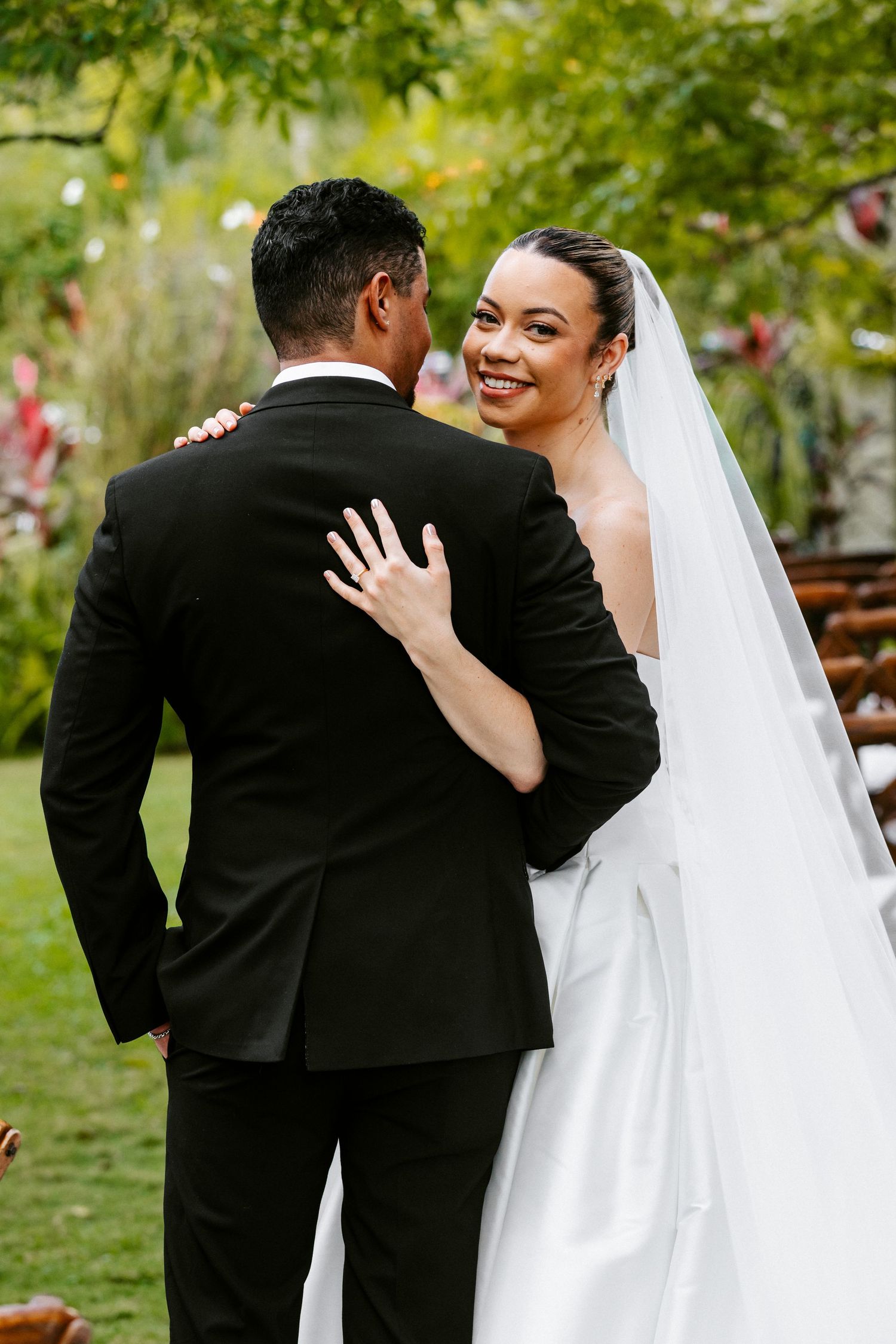 A radiant bride in a white gown and veil smiles over her groom's shoulder during their outdoor wedding.