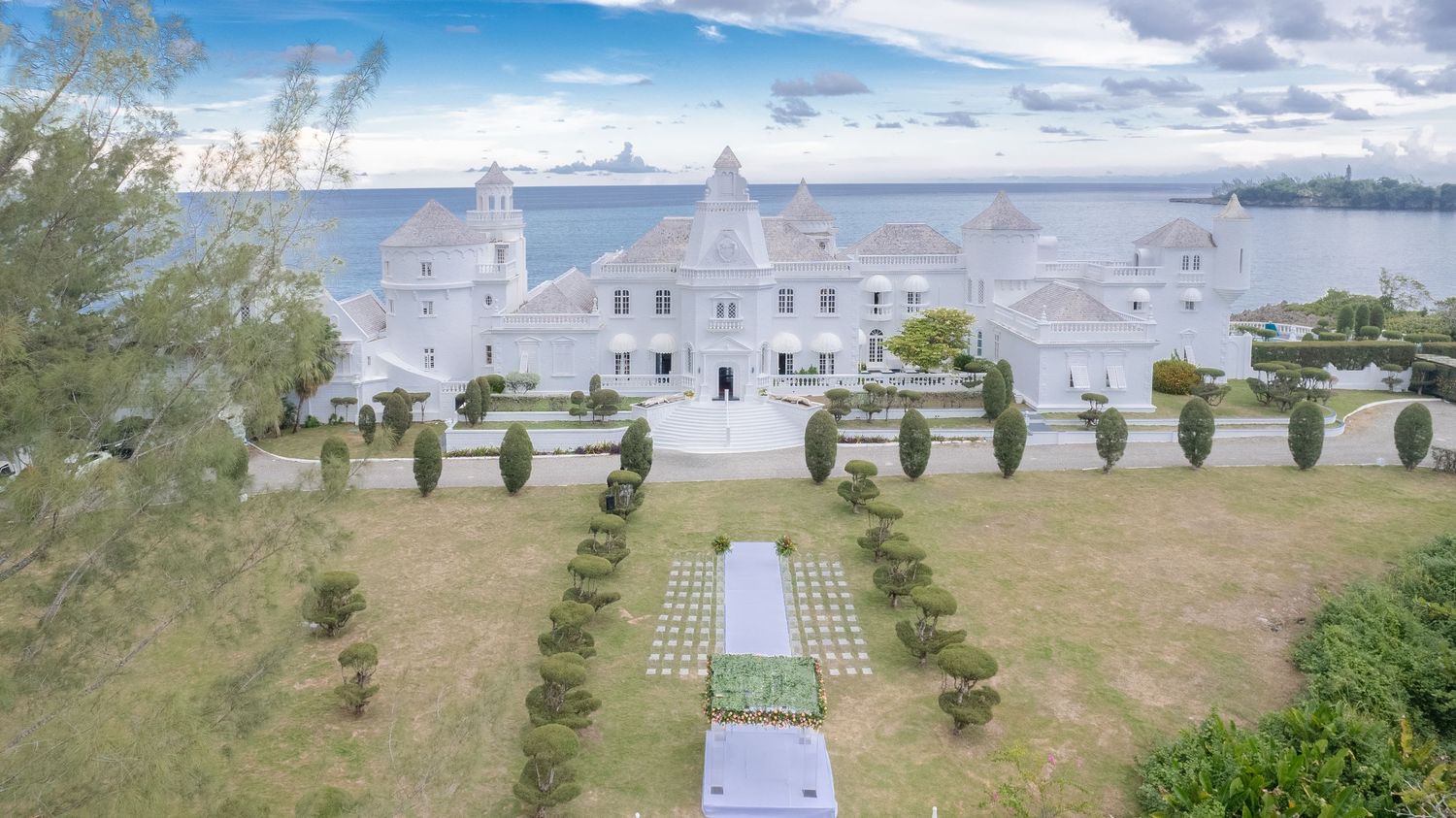 Aerial view of grand white mansion wedding venue with manicured gardens and ocean backdrop in Jamaica.