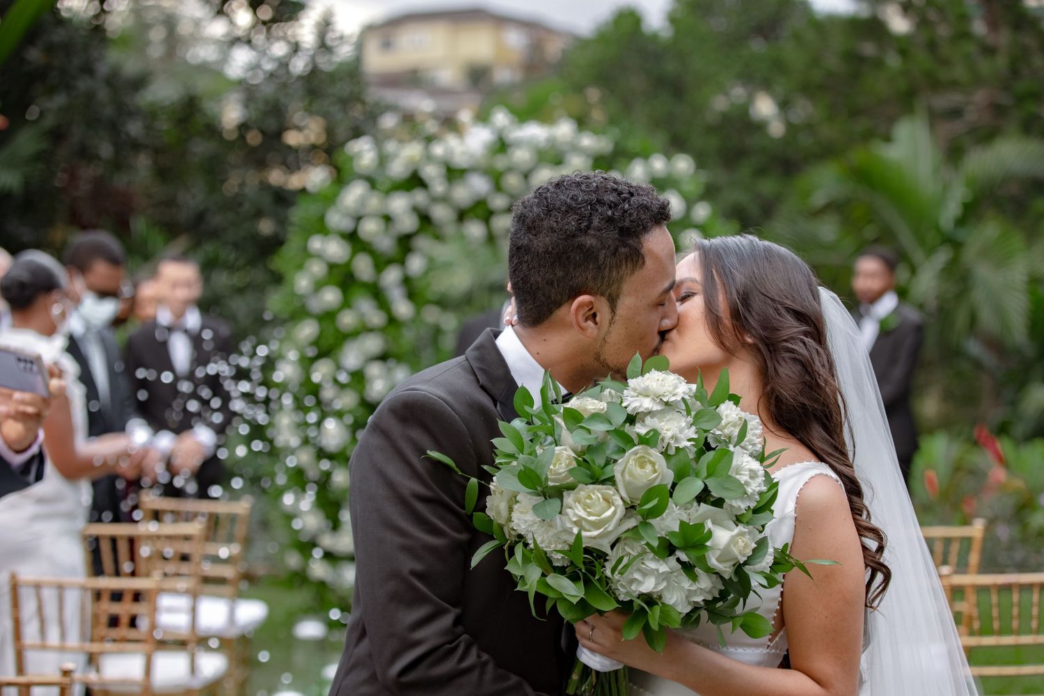 Newlyweds share first kiss at outdoor wedding altar surrounded by white roses and greenery.