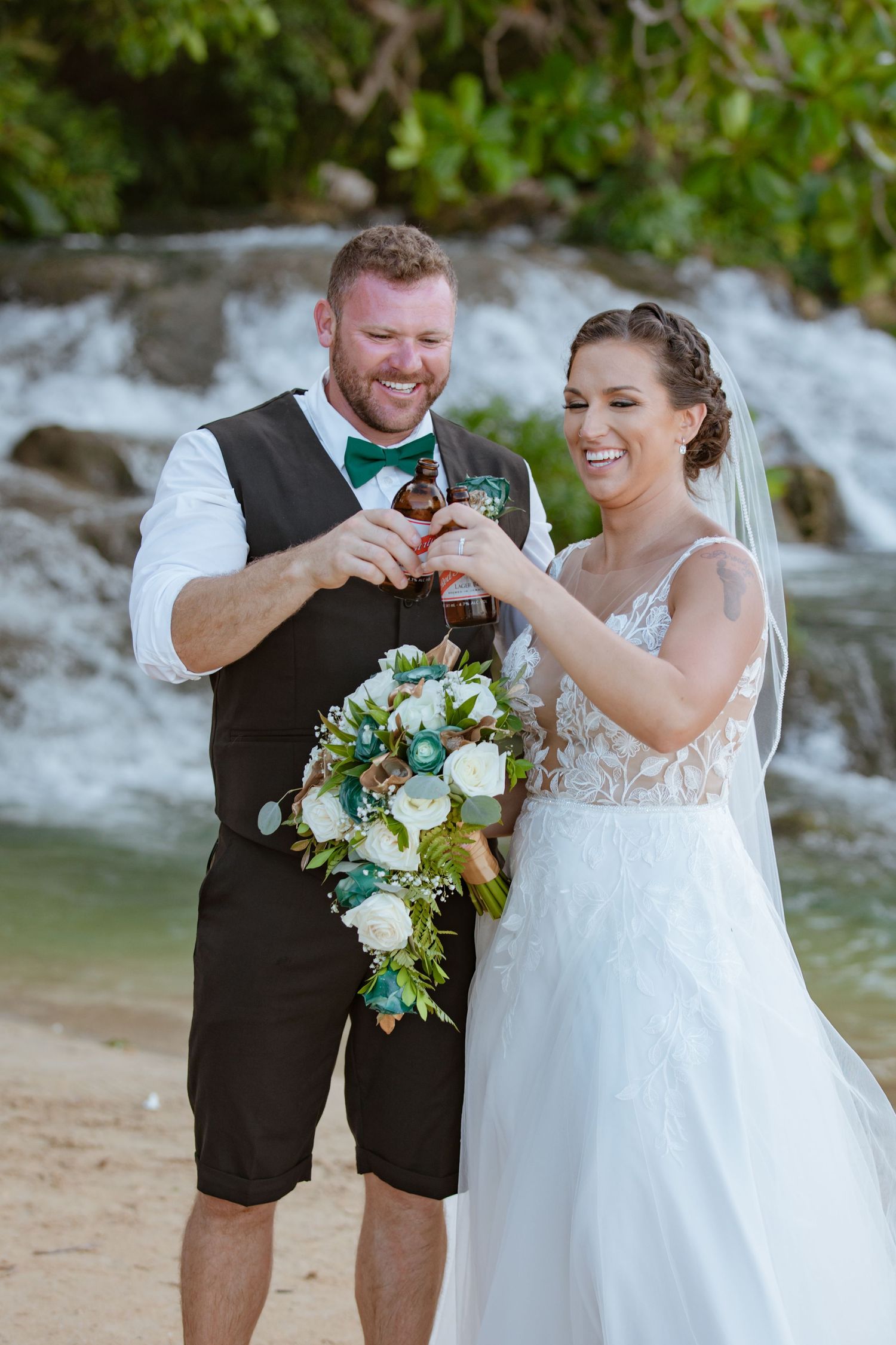 Laughing bride and groom clinking beer bottles together near a lush tropical waterfall.