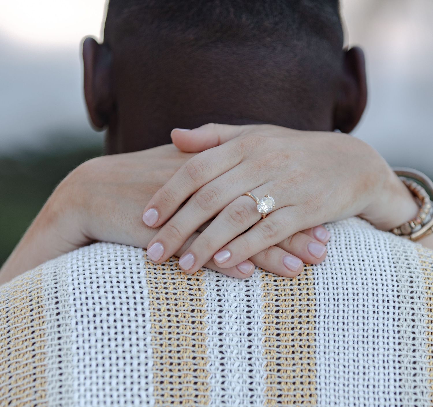 Close-up of womans hand with diamond engagement ring resting on mans striped shirt shoulder.