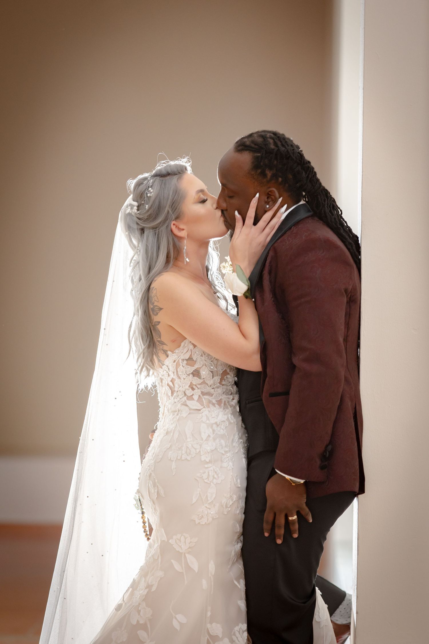 Bride in lace gown and groom in burgundy suit share a tender kiss against a neutral wall.
