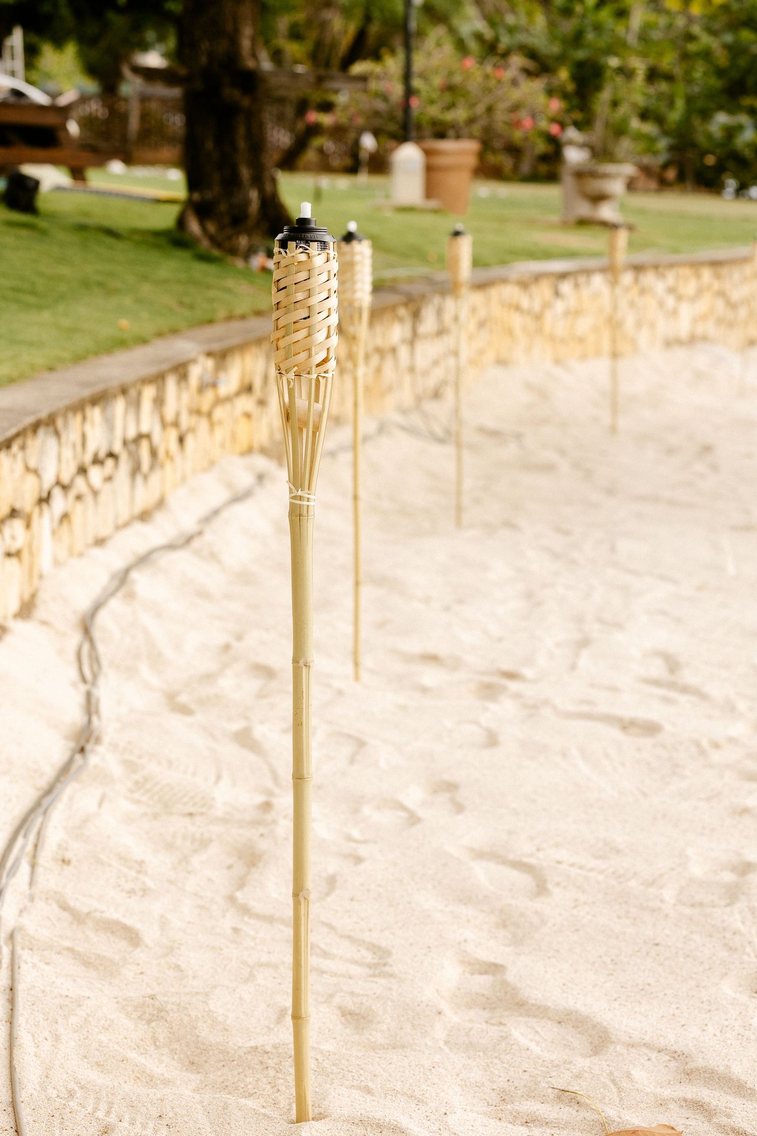 Bamboo tiki torches planted in white sand along a stone wall in a tropical garden setting.