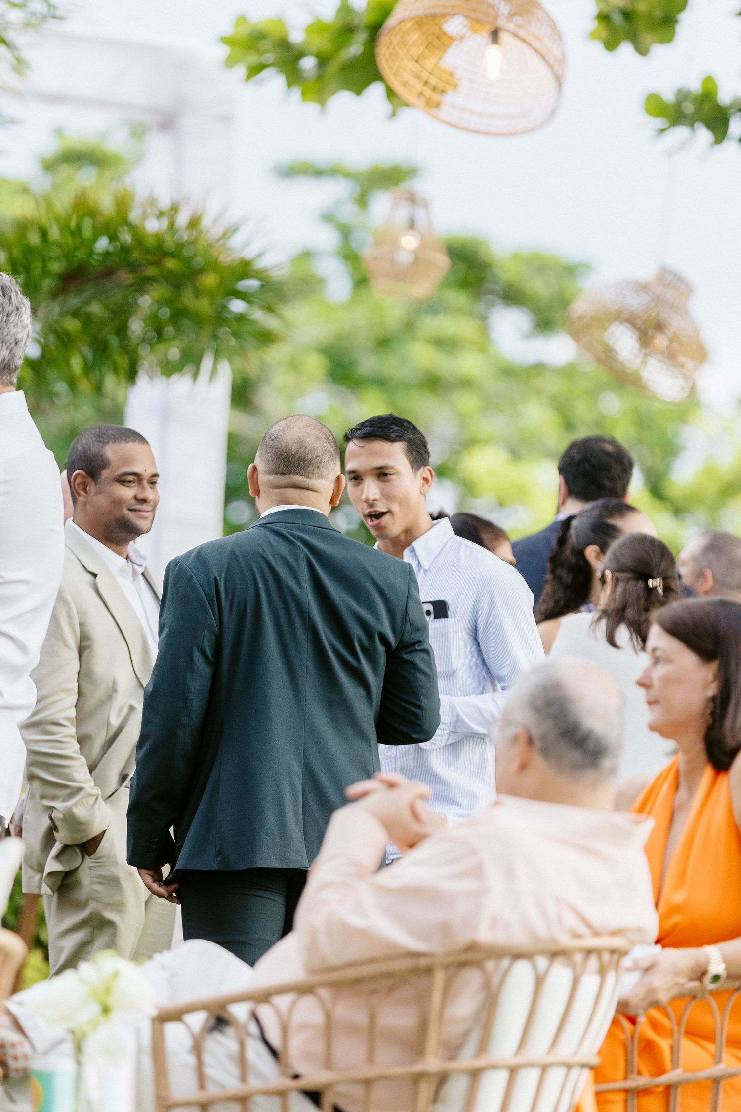 Guests mingling at an outdoor wedding reception, dressed in formal attire, surrounded by lush greenery and warm lighting.