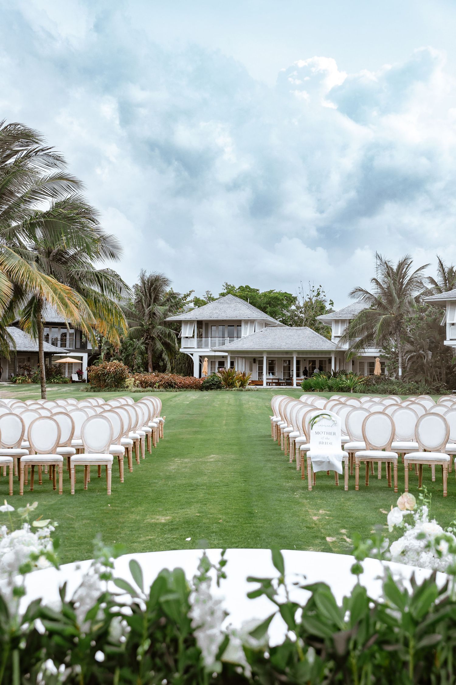 Elegant outdoor wedding ceremony setup with white chairs on green lawn, palm trees, and a tropical villa backdrop.
