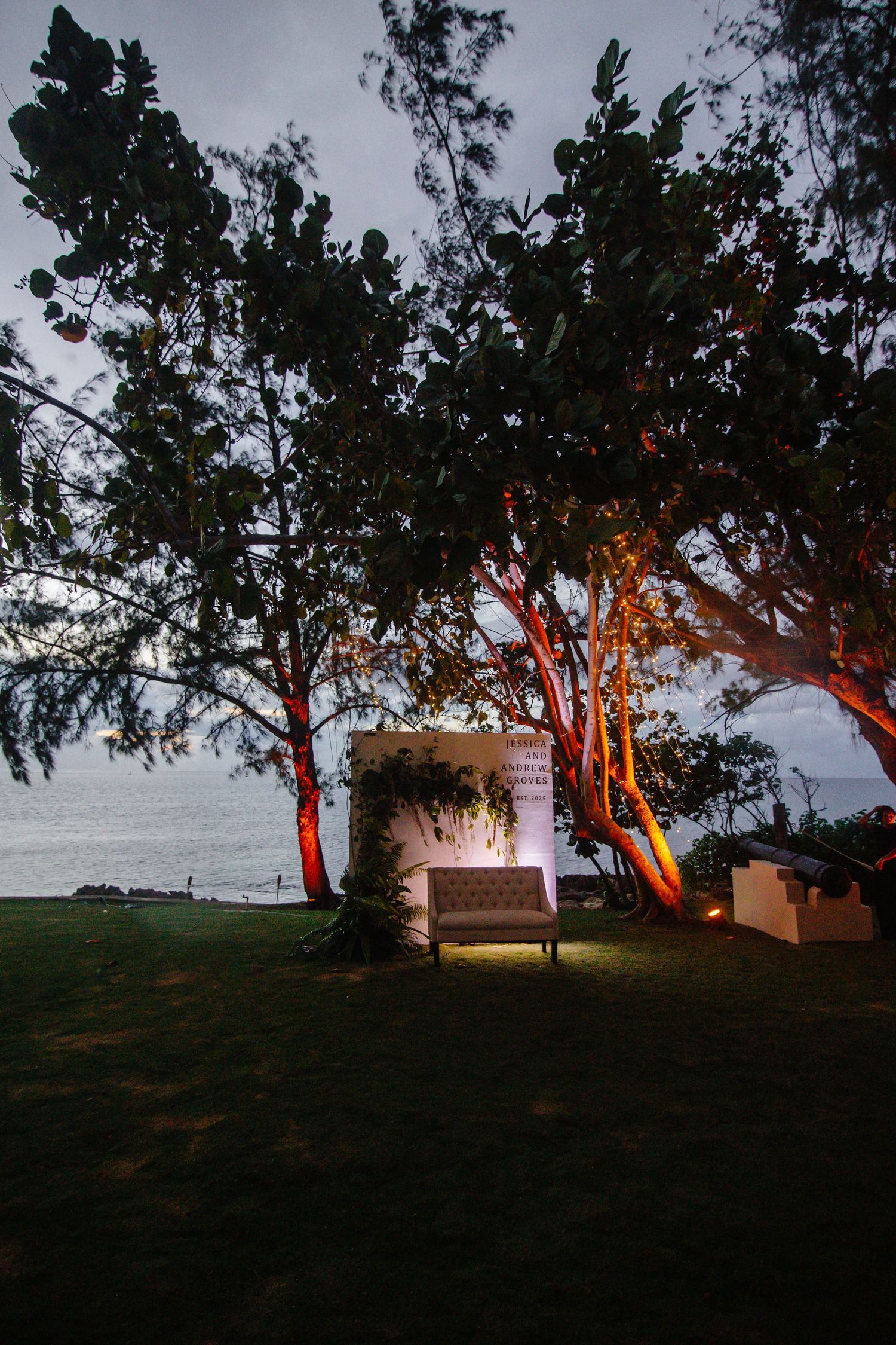 Elegant outdoor wedding setup with illuminated sofa and floral backdrop under large trees by the ocean at dusk.