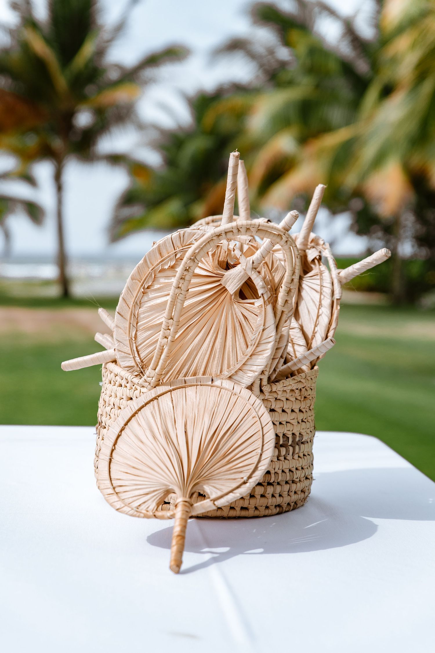 Woven straw basket filled with natural palm leaf hand fans on a white table with tropical palm trees in the background.