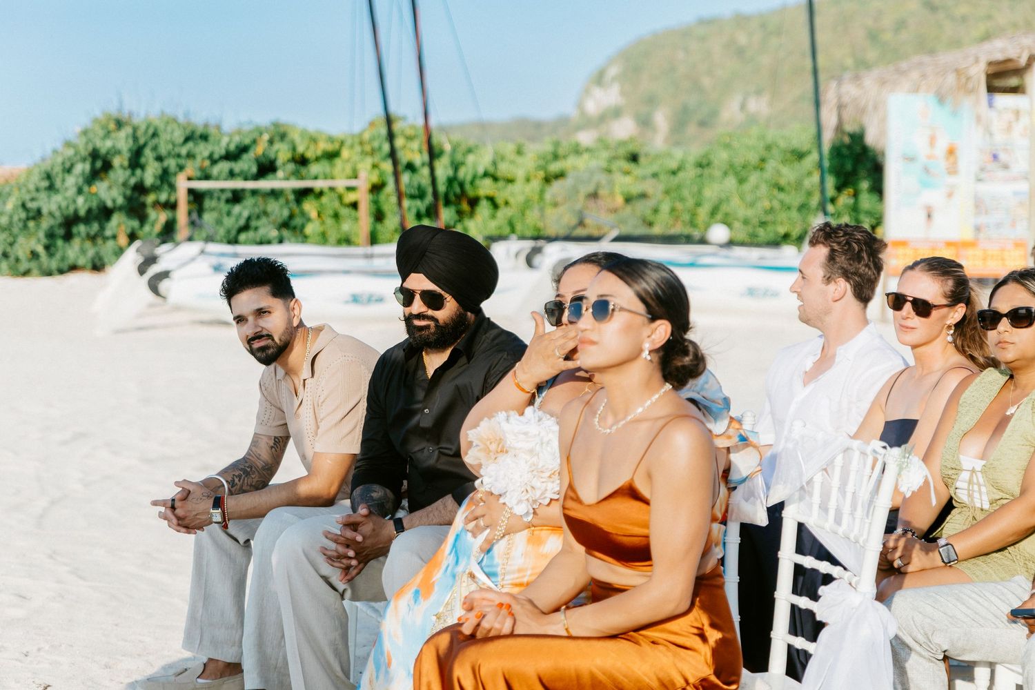 Guests in colorful attire seated on a sunny beach during an outdoor wedding ceremony.