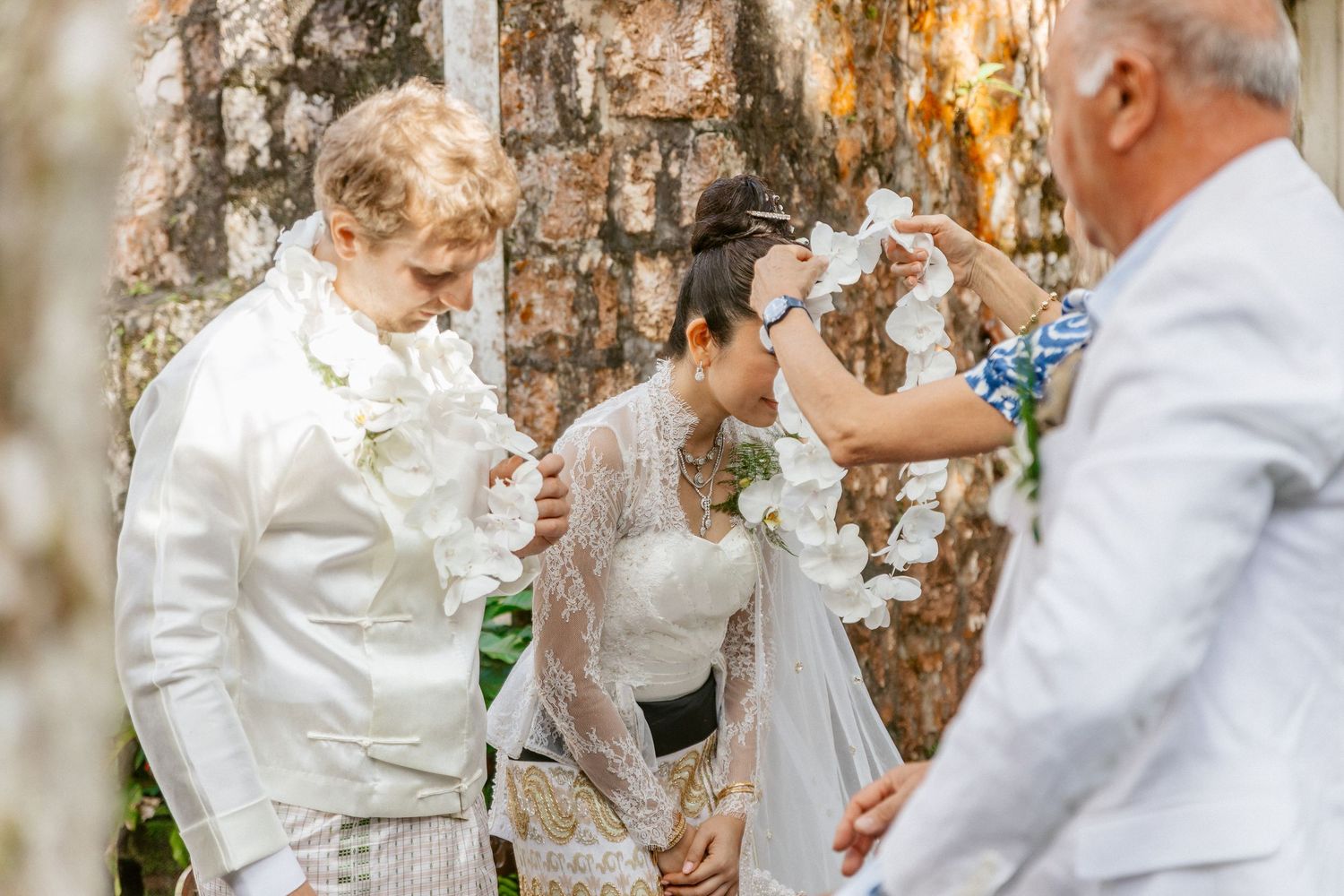 Couple in white wedding attire holding flowers during an outdoor ceremony with an officiant nearby.