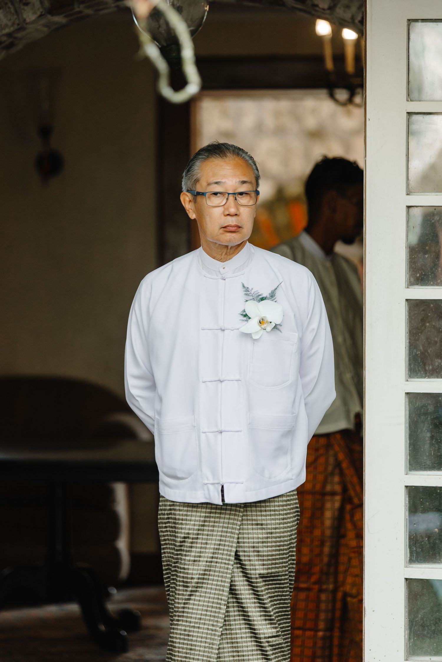 Elderly Asian man in white traditional Chinese shirt with floral boutonniere standing at doorway.