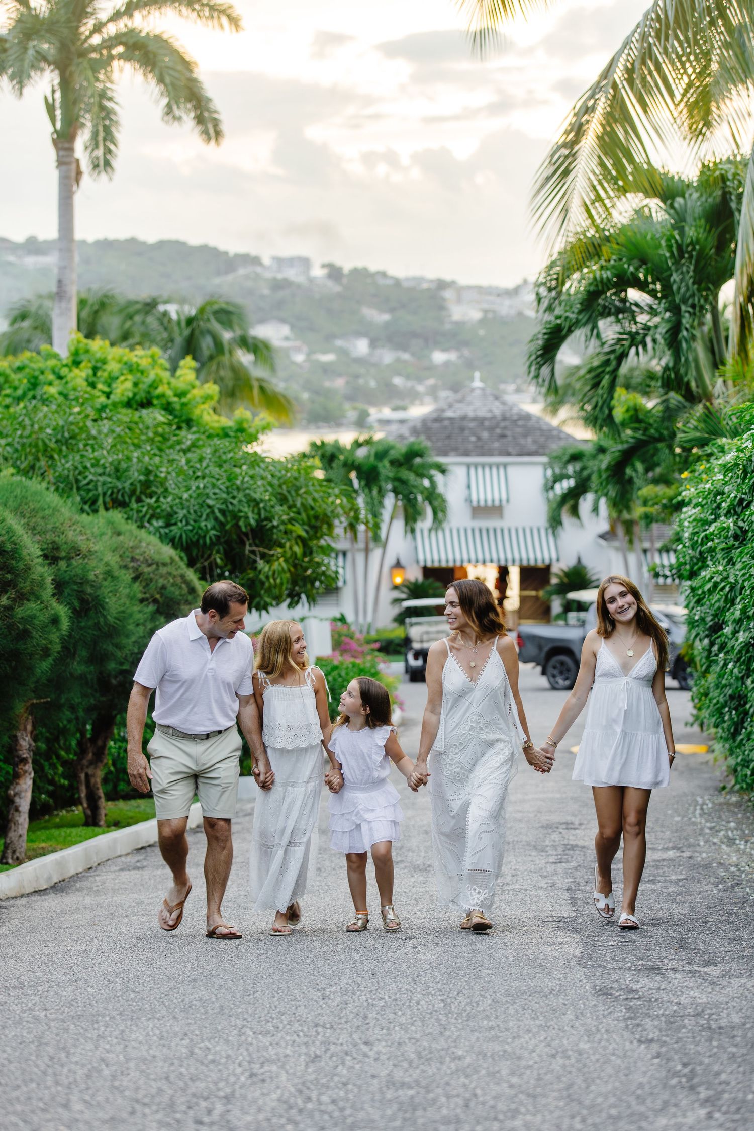Family of five in white outfits walking together on a tropical resort path surrounded by palm trees.