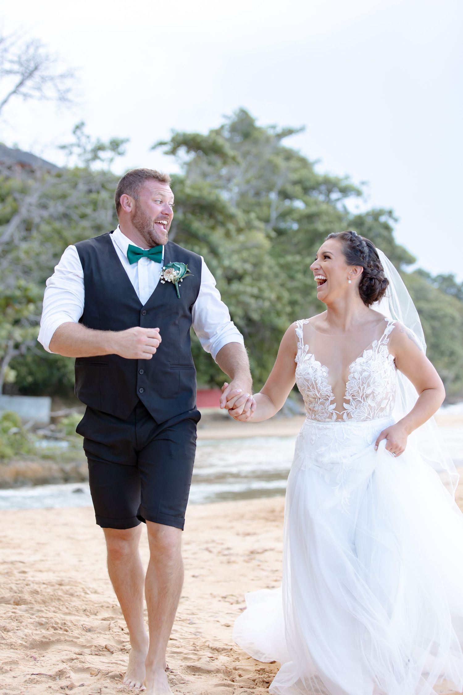 Laughing newlyweds running hand in hand along a tropical beach in wedding attire.