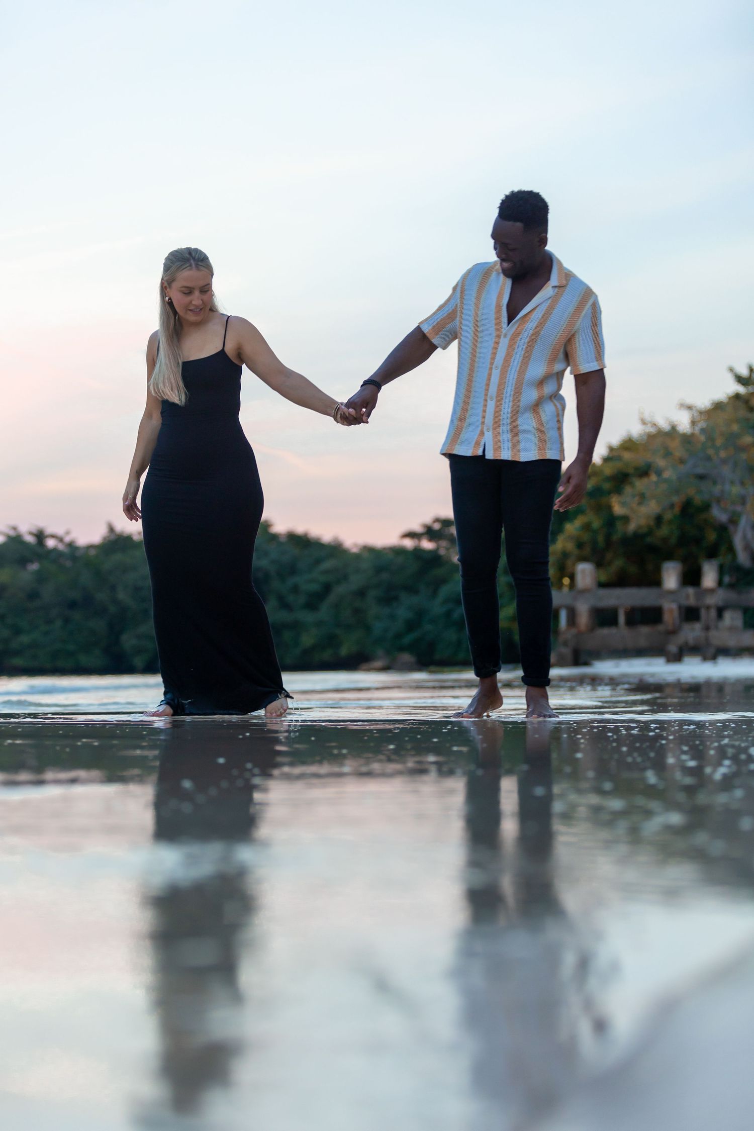 Interracial couple holding hands while walking barefoot on a reflective beach at sunset.