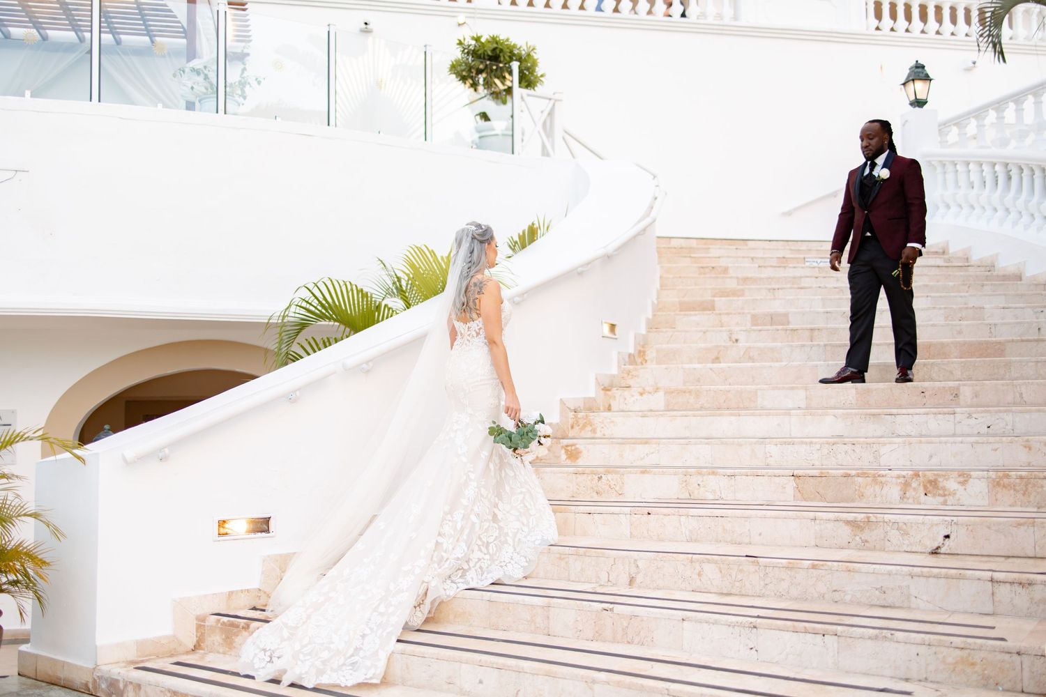 Bride in cathedral veil and groom in burgundy suit on sweeping marble staircase at resort.