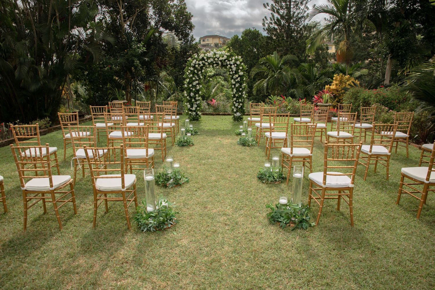Elegant outdoor wedding ceremony setup with gold chiavari chairs and white floral arch on lawn.
