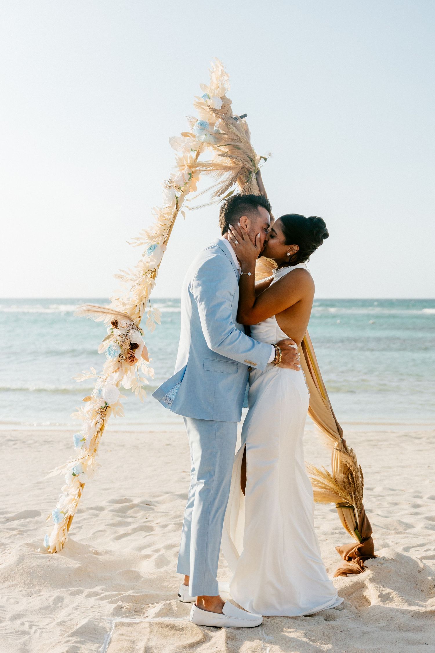 Couple kissing at beach wedding ceremony under boho arch with pampas grass decorations.